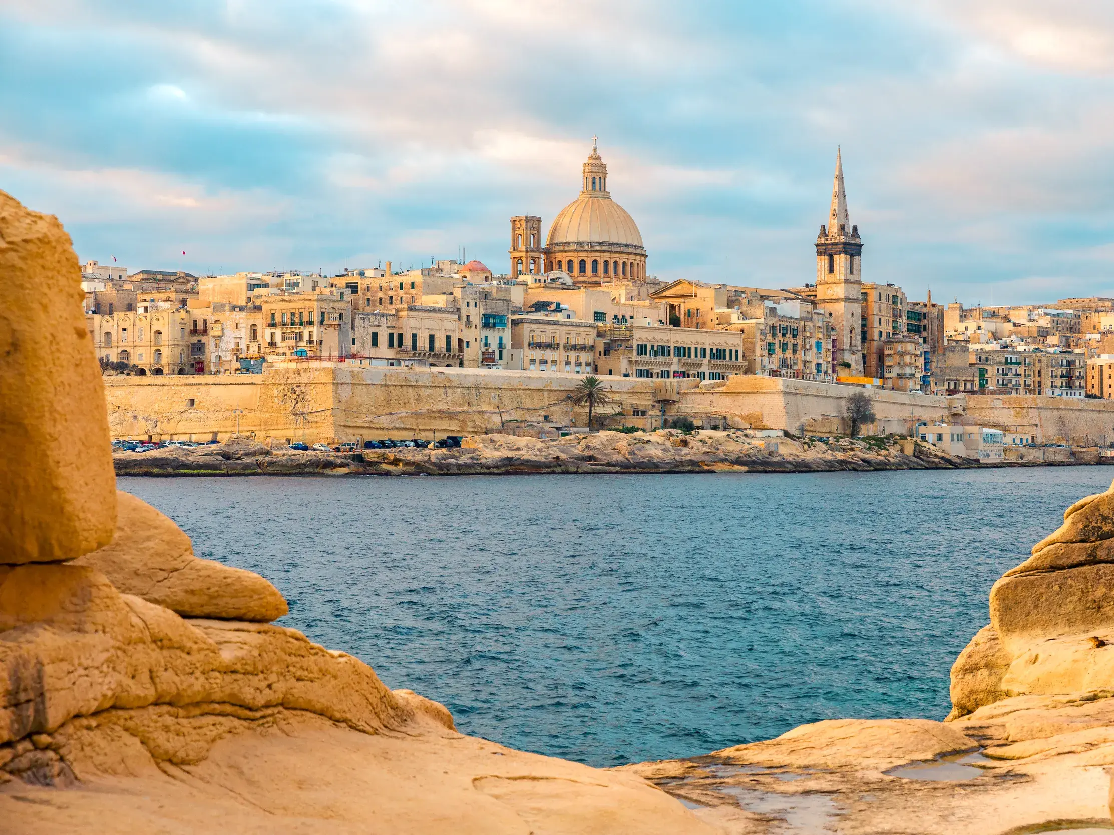 Von Felsen gerahmter Blick auf die Skyline der Ritterstadt Valletta in Malta mit ihrer Kuppel und den Festungsmauern am Meer.