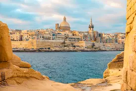 Von Felsen gerahmter Blick auf die Skyline der Ritterstadt Valletta in Malta mit ihrer Kuppel und den Festungsmauern am Meer.