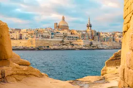 Von Felsen gerahmter Blick auf die Skyline der Ritterstadt Valletta in Malta mit ihrer Kuppel und den Festungsmauern am Meer.