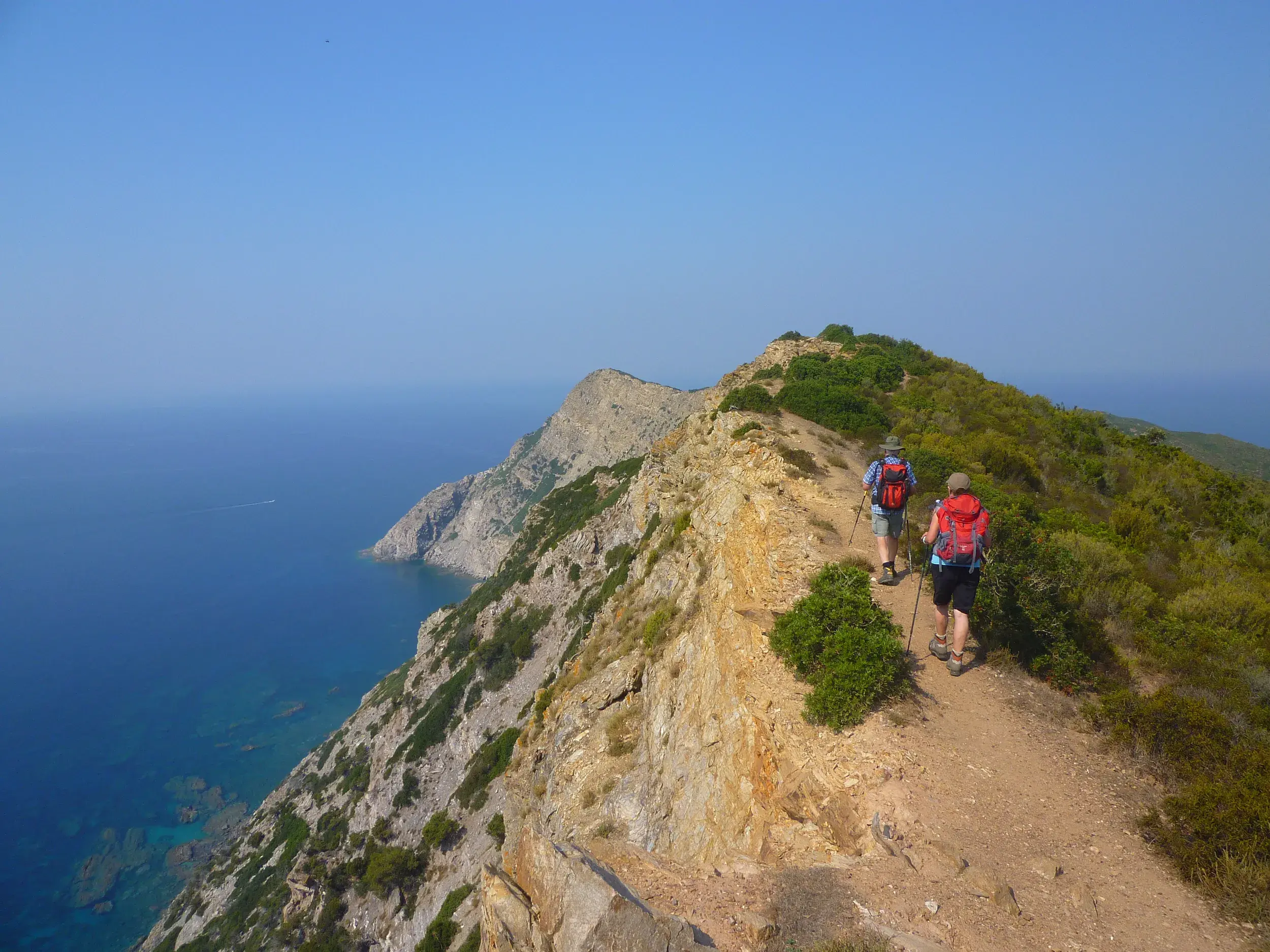 Zwei Wanderer mit Rucksäcken auf einem aussichtsreichen Küstenpfad entlang der Steilküste Sardiniens bei Sonnenschein.