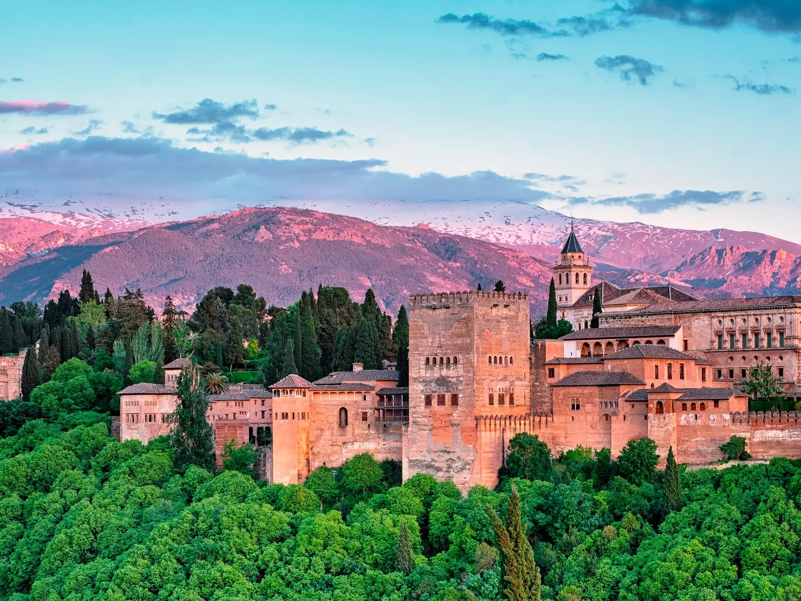 Die maurische Palaststadt Alhambra in Granada, umgeben von Bäumen, vor der im Abendlicht leuchtenden Sierra Nevada.