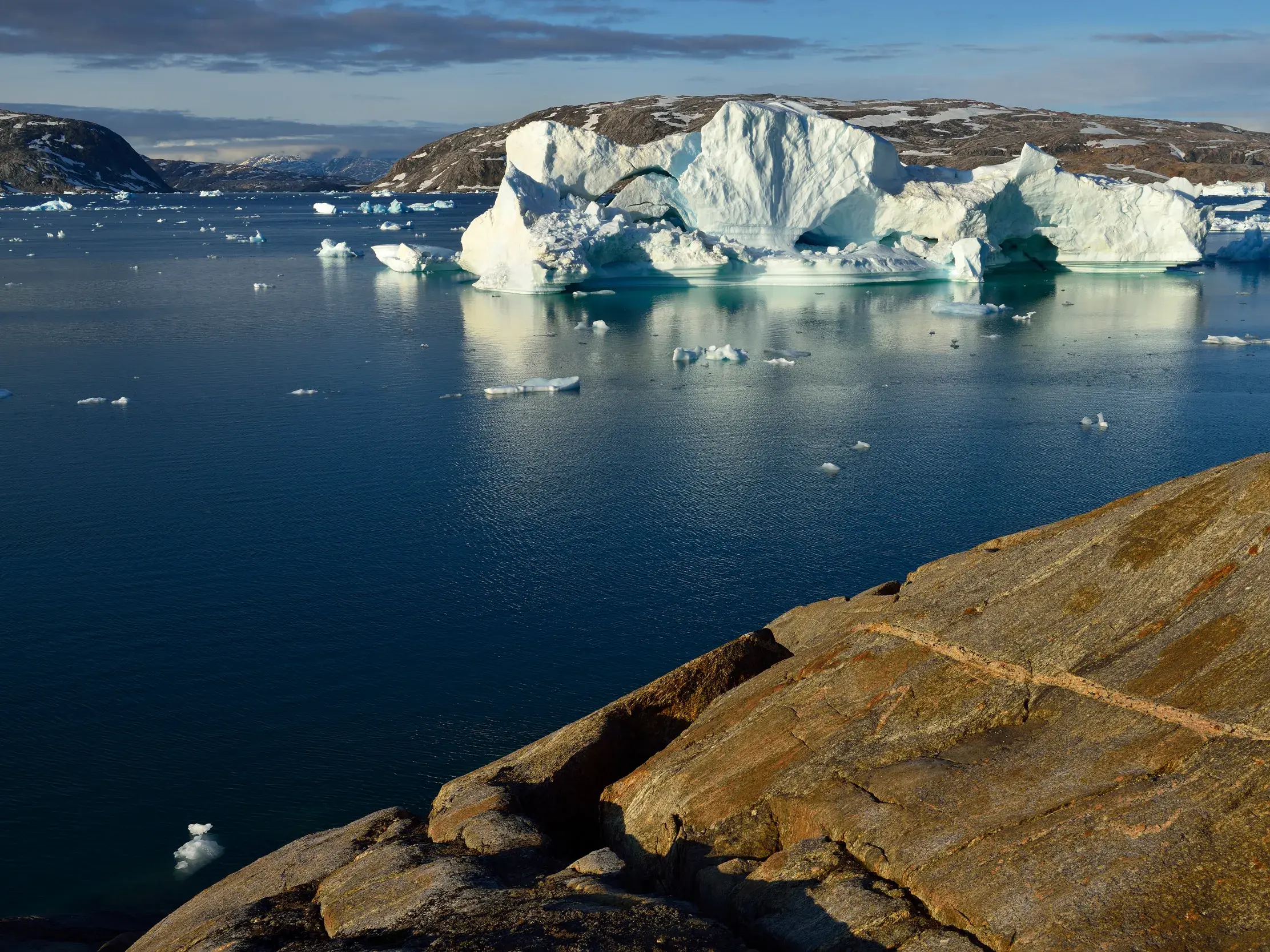 Gigantischer Eisberg treibt im tiefblauen Wasser des Sermilik-Fjords in Ostgrönland, gesehen von der felsigen Küste.