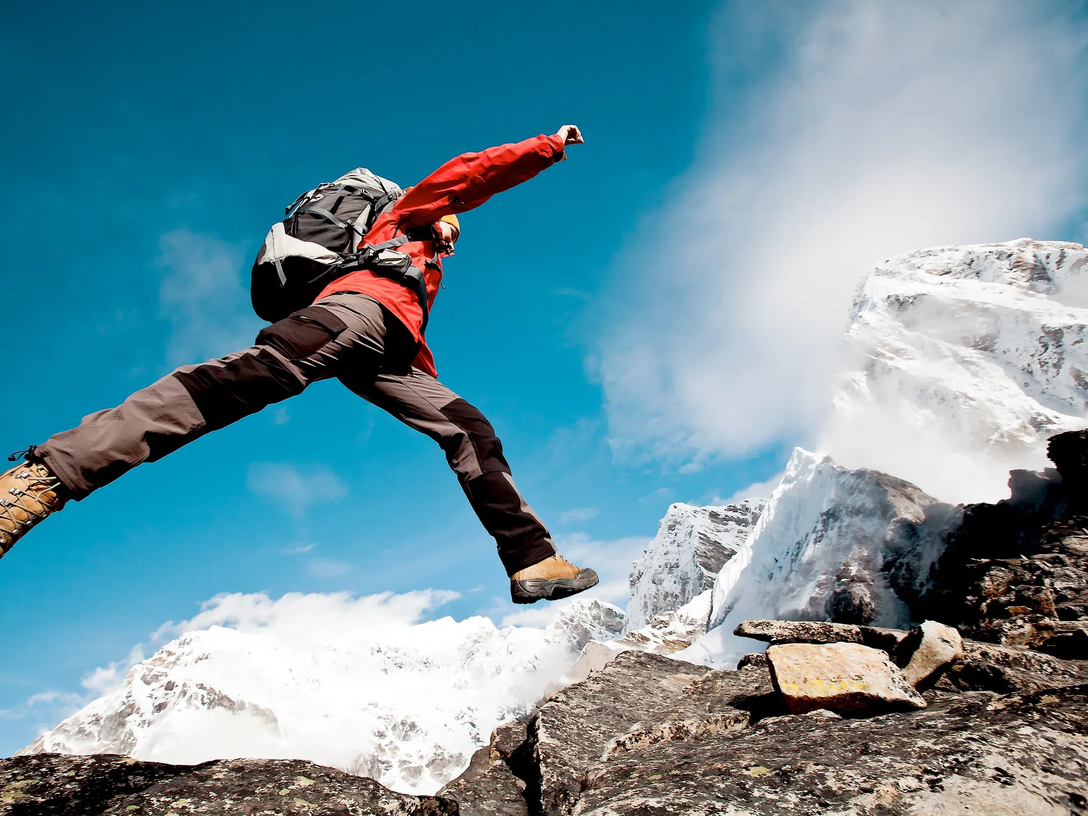 Ein Wanderer in roter Jacke springt über Felsen vor den majestätischen, schneebedeckten Gipfeln des Himalaya in Nepal.