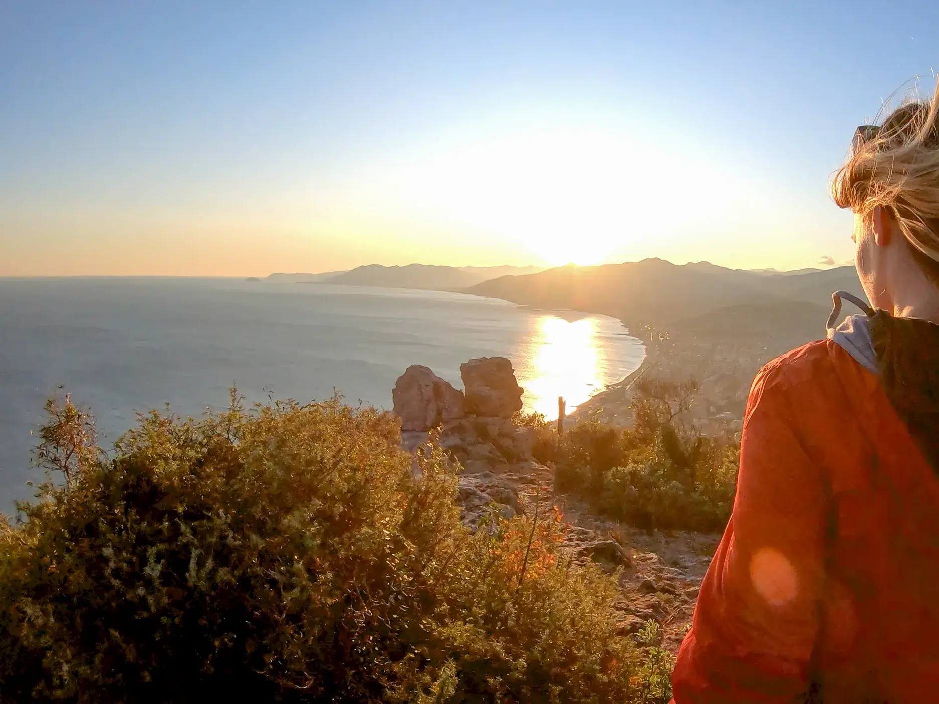 Eine Frau blickt bei einer Küstenwanderung von einem Aussichtspunkt auf die im Sonnenuntergang glitzernde ligurische Küste.