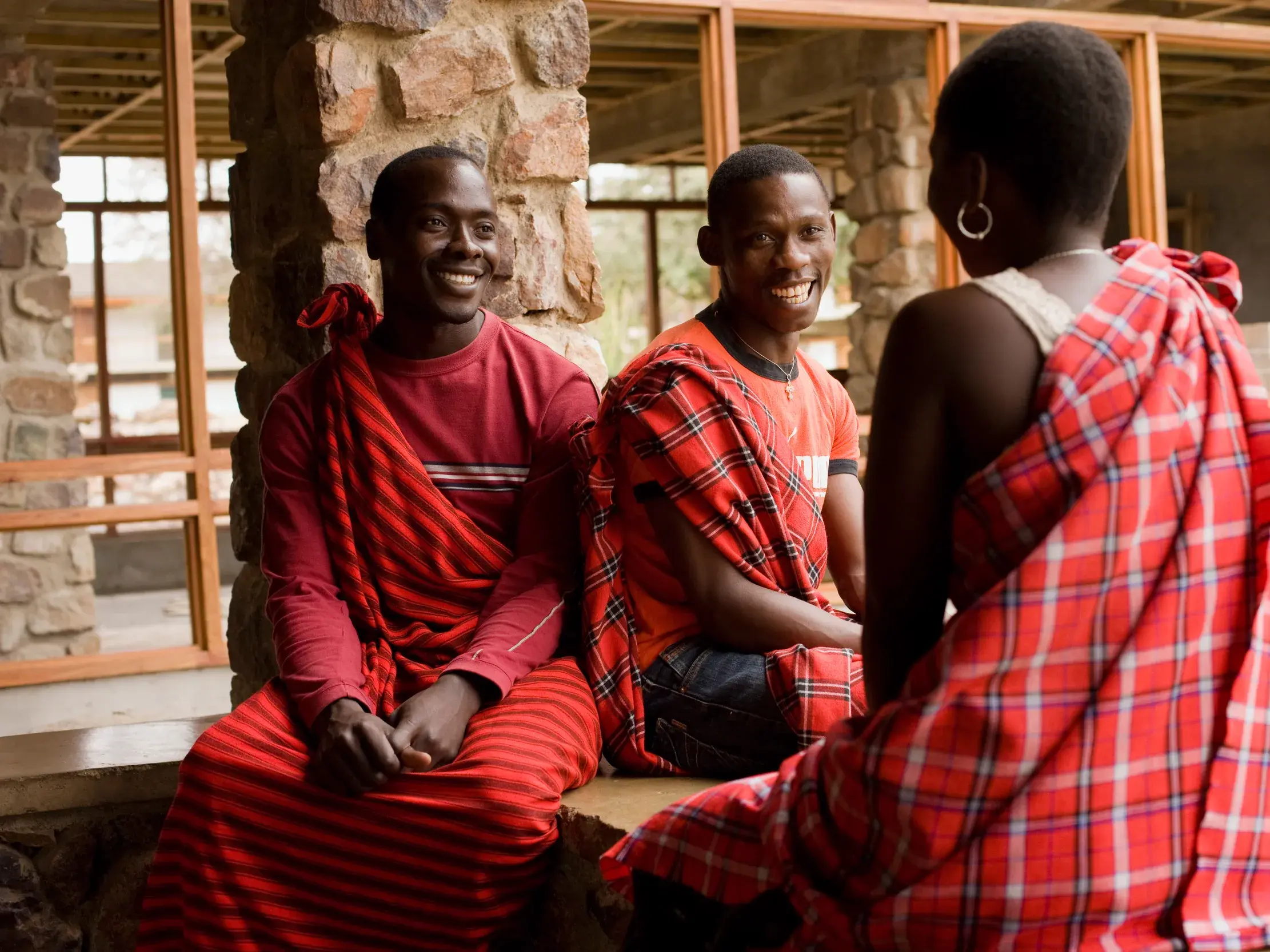 Zwei lächelnde Massai-Männer im Gespräch mit einer Frau, alle in traditionellen roten Shuka-Tüchern, in einer Lodge in Tansania.