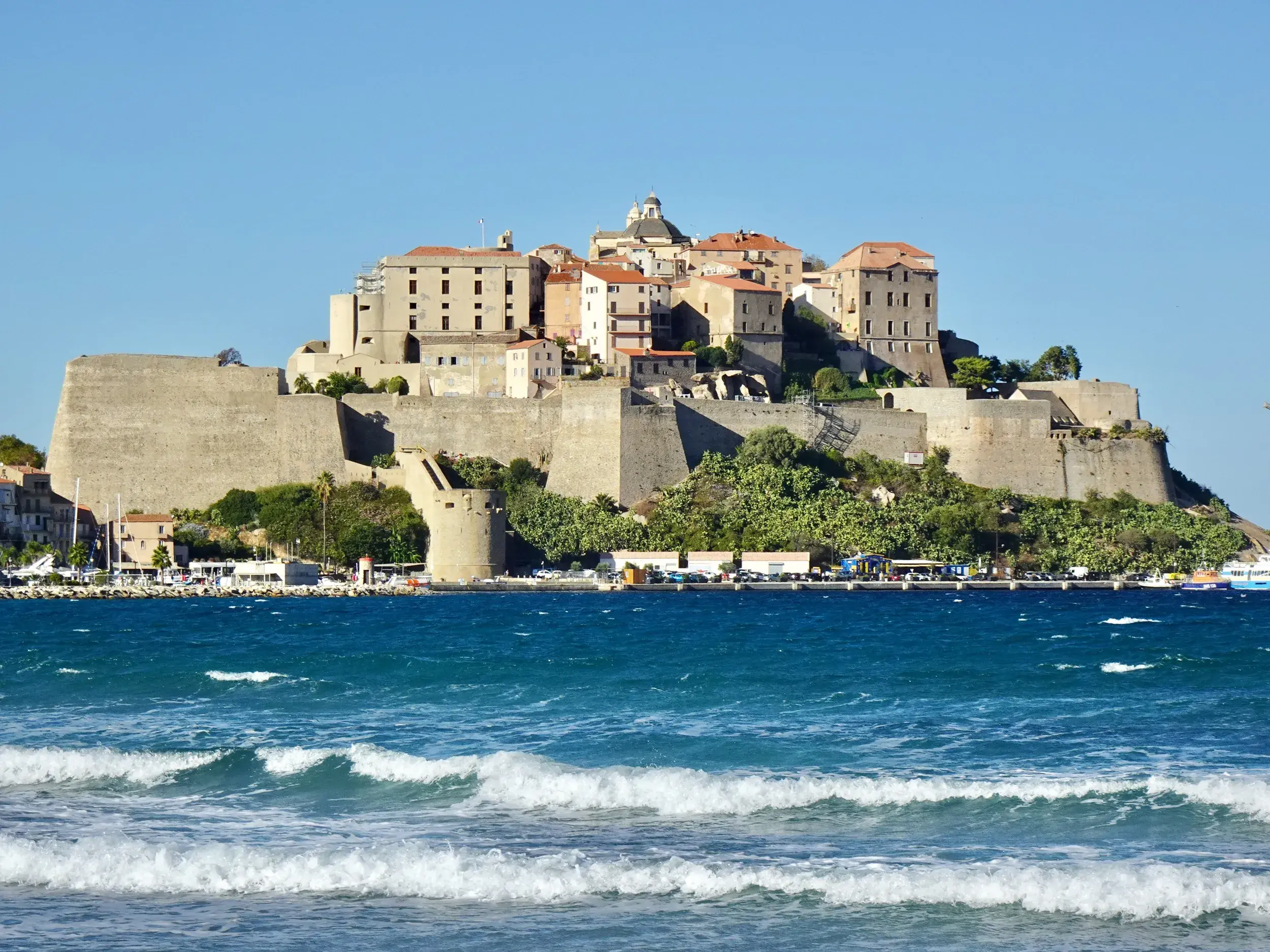 Sonnige Küstenansicht der Zitadelle von Calvi auf Korsika mit dem türkisen Mittelmeer und weißen Wellen im Vordergrund.