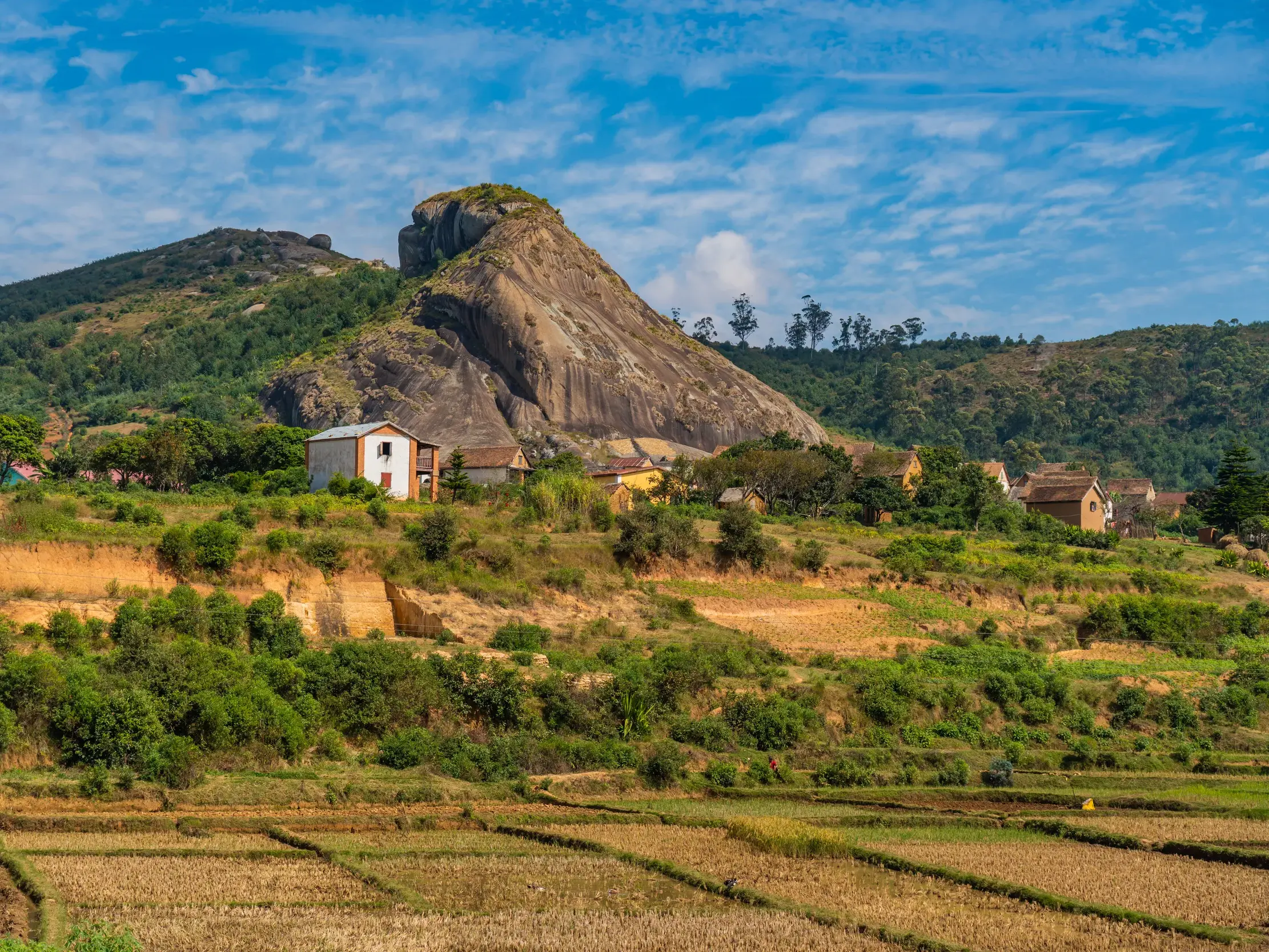 Hügelige Landschaft im Hochland von Madagaskar mit terrassierten Reisfeldern, einem Dorf und einer großen Felsformation.