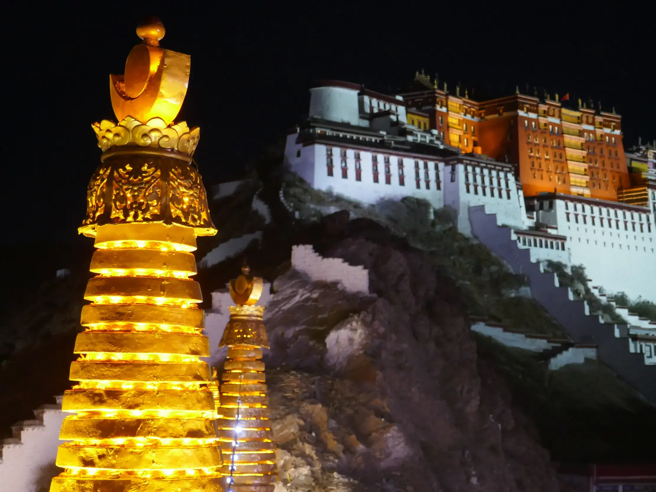 Goldene, illuminierte Chörten im Vordergrund vor dem nächtlich angestrahlten Potala-Palast in Lhasa während einer Trekkingreise.