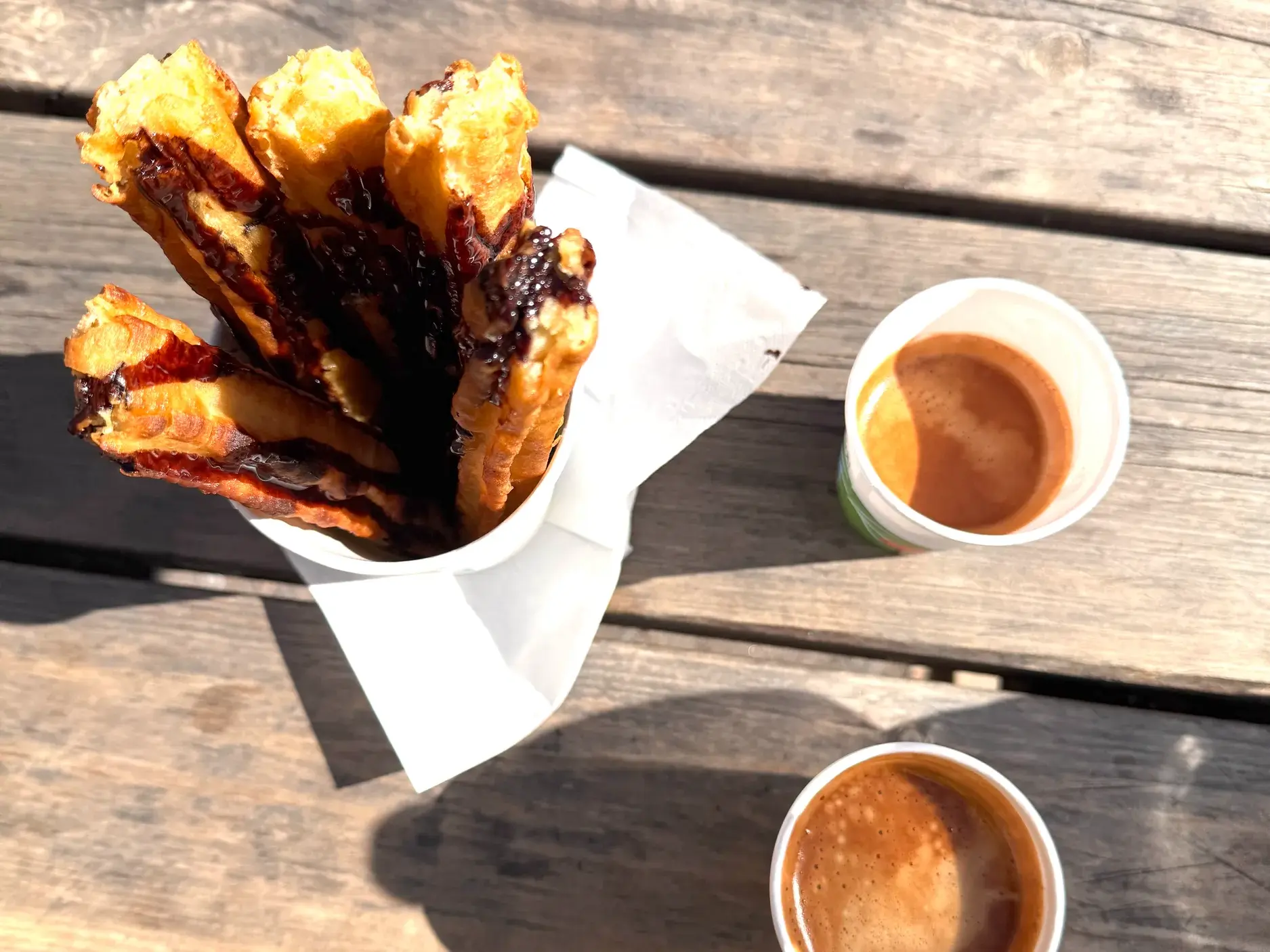 Typisch spanische Churros mit heißer Schokolade in einer Papiertüte, serviert mit einem Kaffee auf einem sonnigen Holztisch.