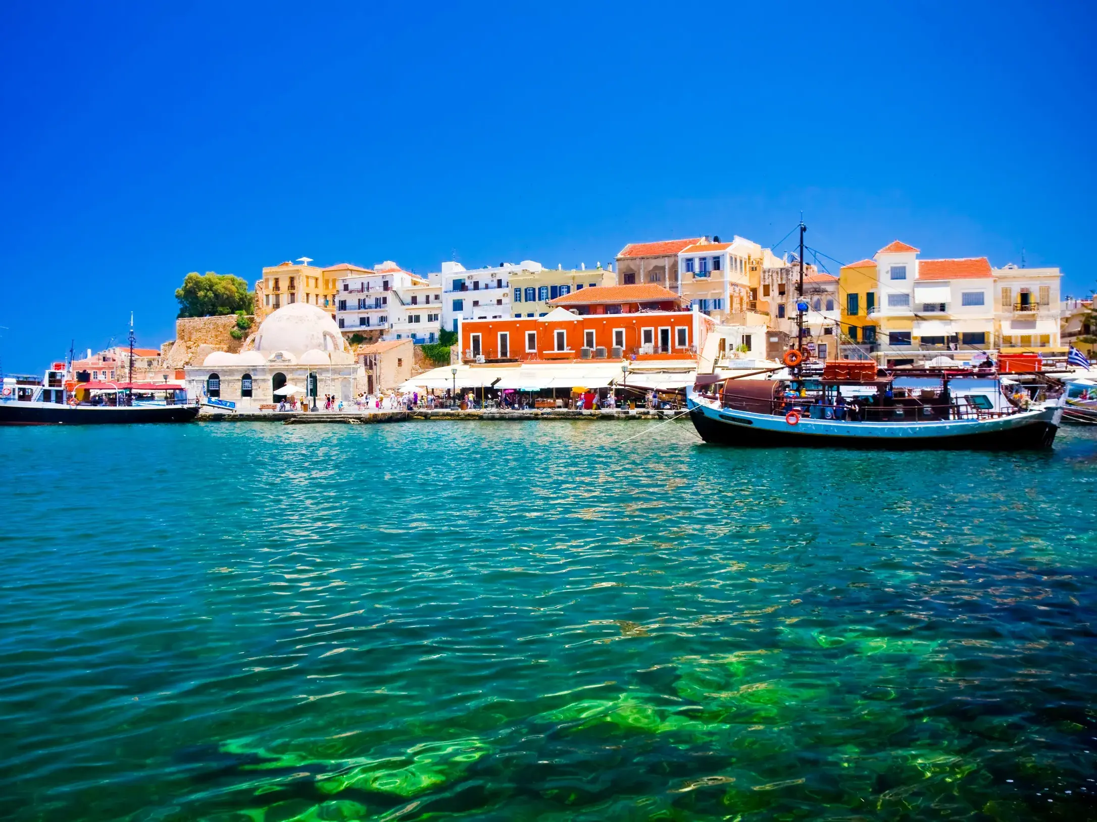 Bunte Hafenpromenade von Chania mit der Hasan-Pascha-Moschee und Fischerbooten, gesehen vom türkisfarbenen Meer auf Kreta.