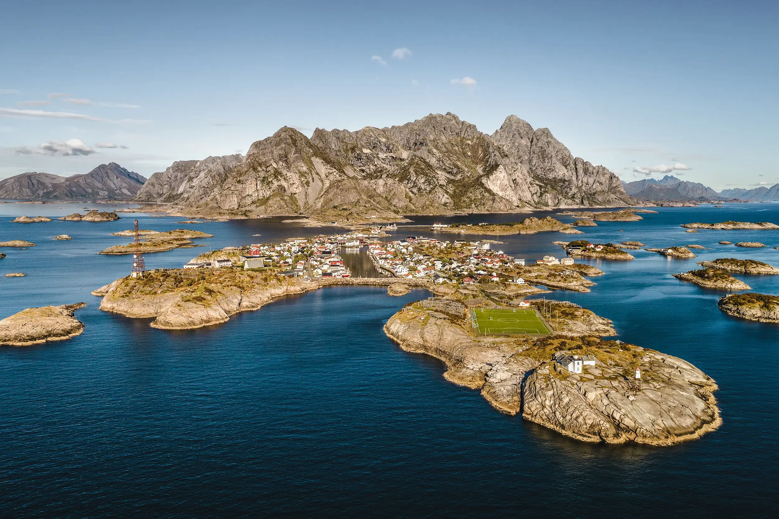 Luftaufnahme des Fischerdorfs Henningsvær auf den Lofoten mit dem Fußballplatz, umgeben von blauem Meer und schroffer Bergkulisse.