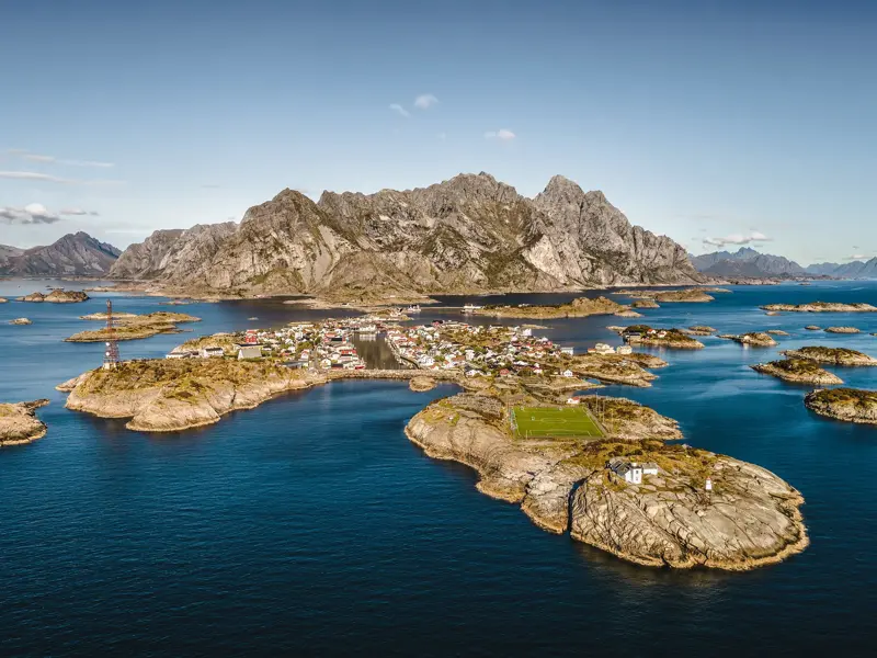 Luftaufnahme des Fischerdorfs Henningsvær auf den Lofoten mit dem Fußballplatz, umgeben von blauem Meer und schroffer Bergkulisse.