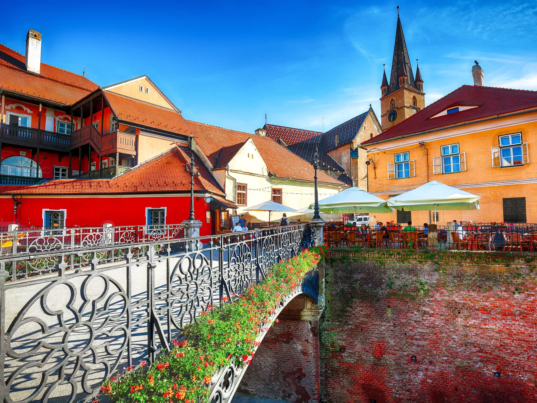 Sonniger Blick über die blumengeschmückte Lügenbrücke in Hermannstadt auf bunte Patrizierhäuser und Cafés in der Altstadt.