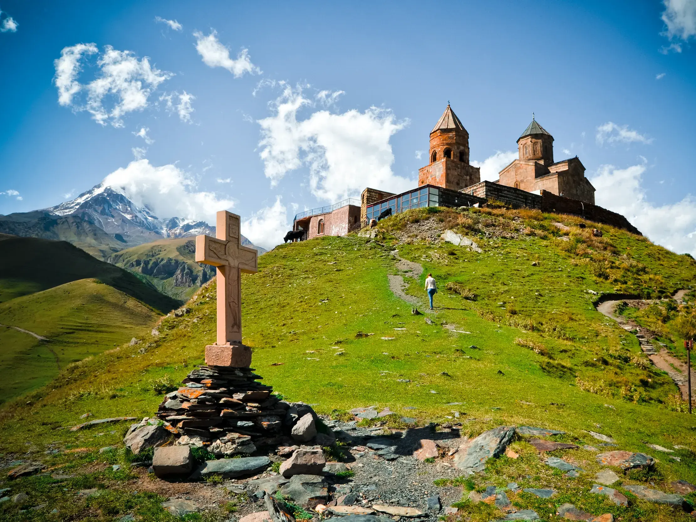 Eine Person wandert auf einem Pfad zur Gergeti-Dreifaltigkeitskirche, die auf einem Hügel vor dem Berg Kasbek in Georgien thront.