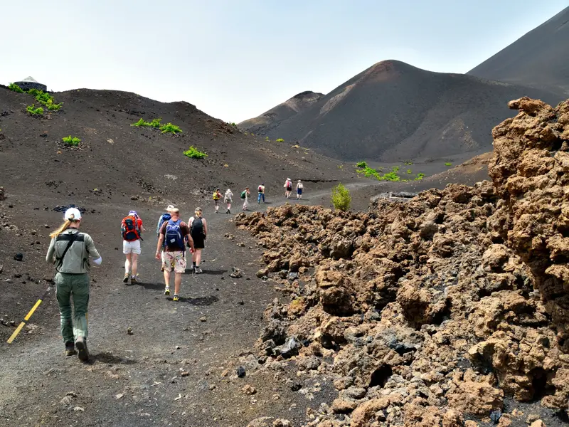 Eine geführte Wandergruppe mit Rucksäcken auf einem Pfad durch die schwarze Vulkanlandschaft der Chã das Caldeiras auf Fogo.