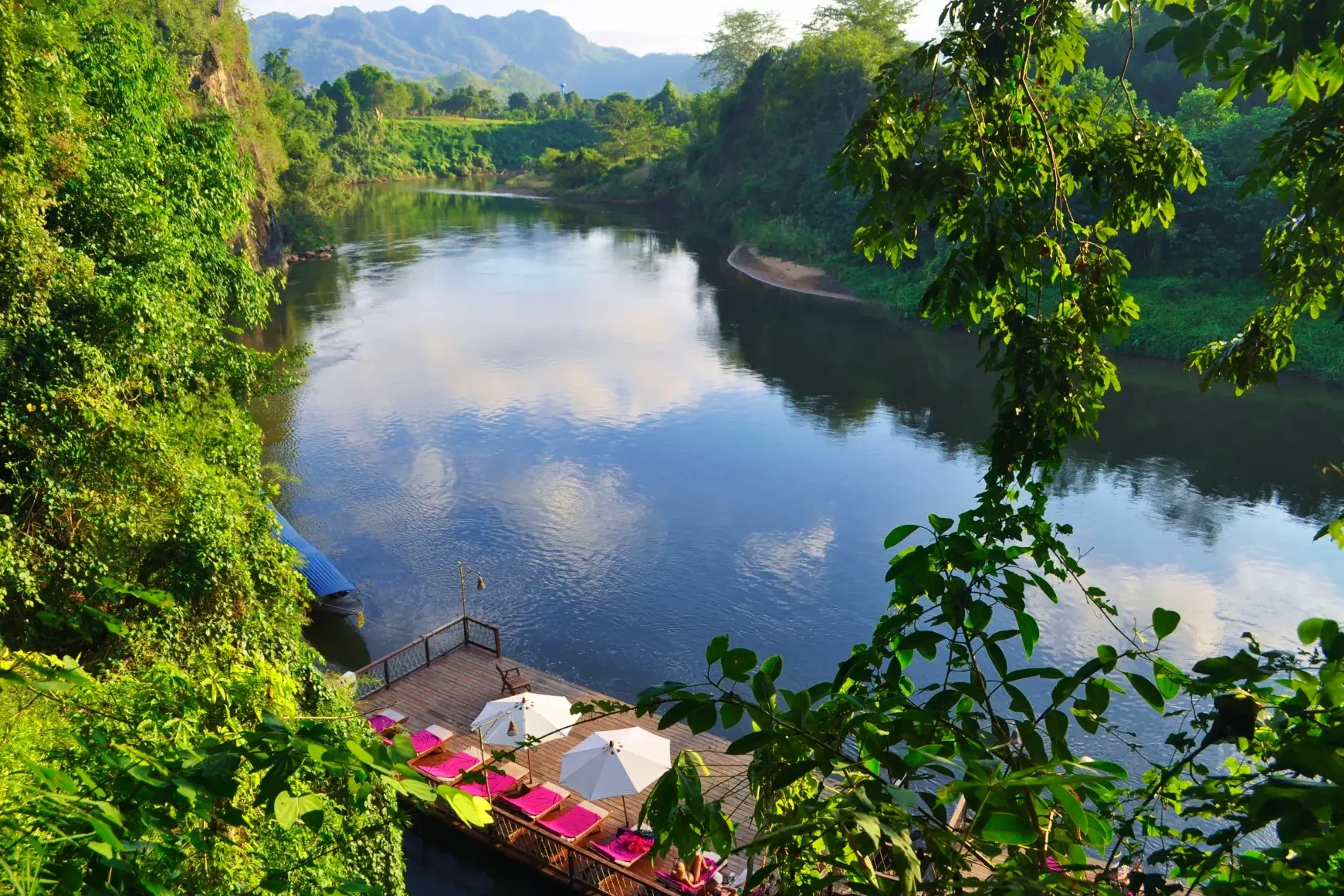 Sonnendeck eines Floßhotels mit Liegestühlen am ruhigen River Kwai, umgeben von tropischem Dschungel in Thailand.