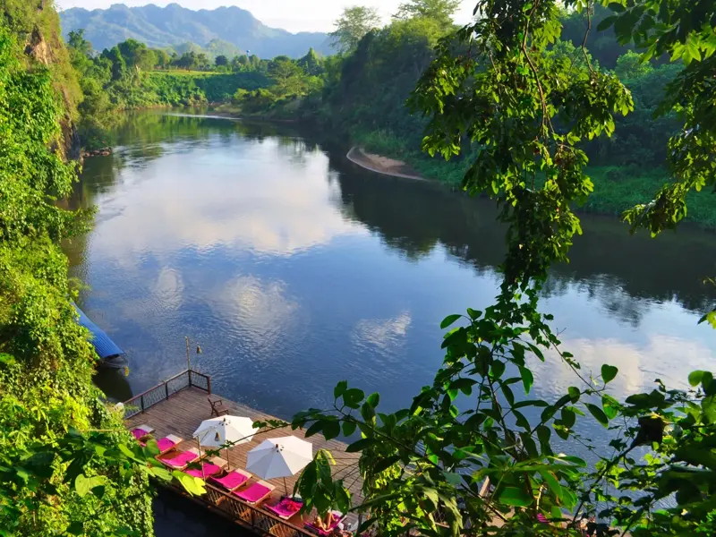 Sonnendeck eines Floßhotels mit Liegestühlen am ruhigen River Kwai, umgeben von tropischem Dschungel in Thailand.