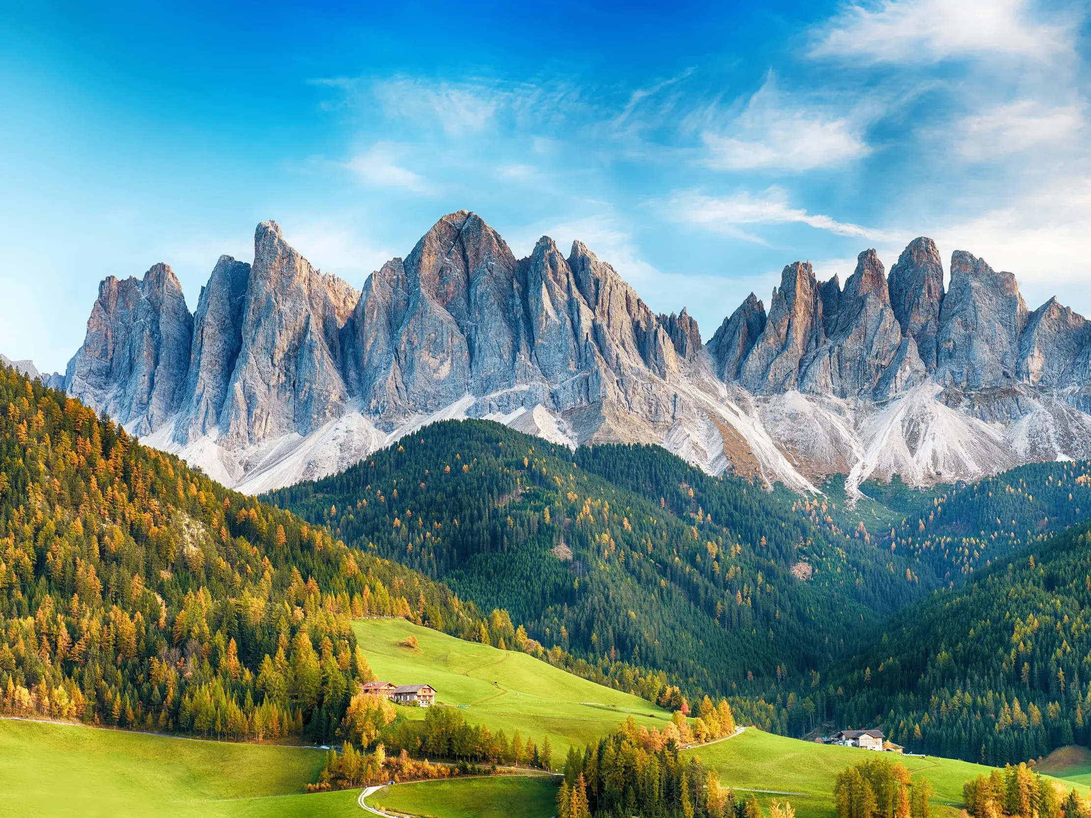 Die spitzen Felsgipfel der Geislergruppe in den Dolomiten über grünen Almwiesen und herbstlichen Wäldern in Südtirol.