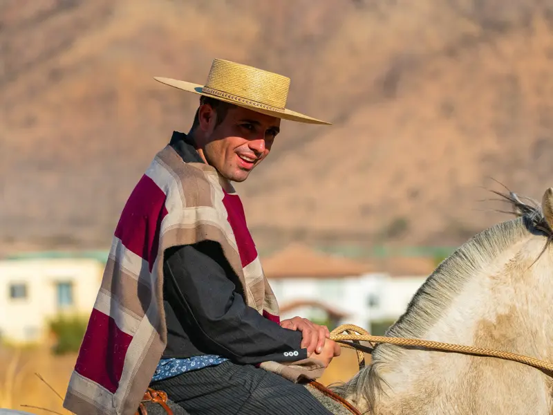 Lächelnder Gaucho mit traditionellem Poncho und Hut reitet auf einem weißen Pferd durch die argentinische Pampa.
