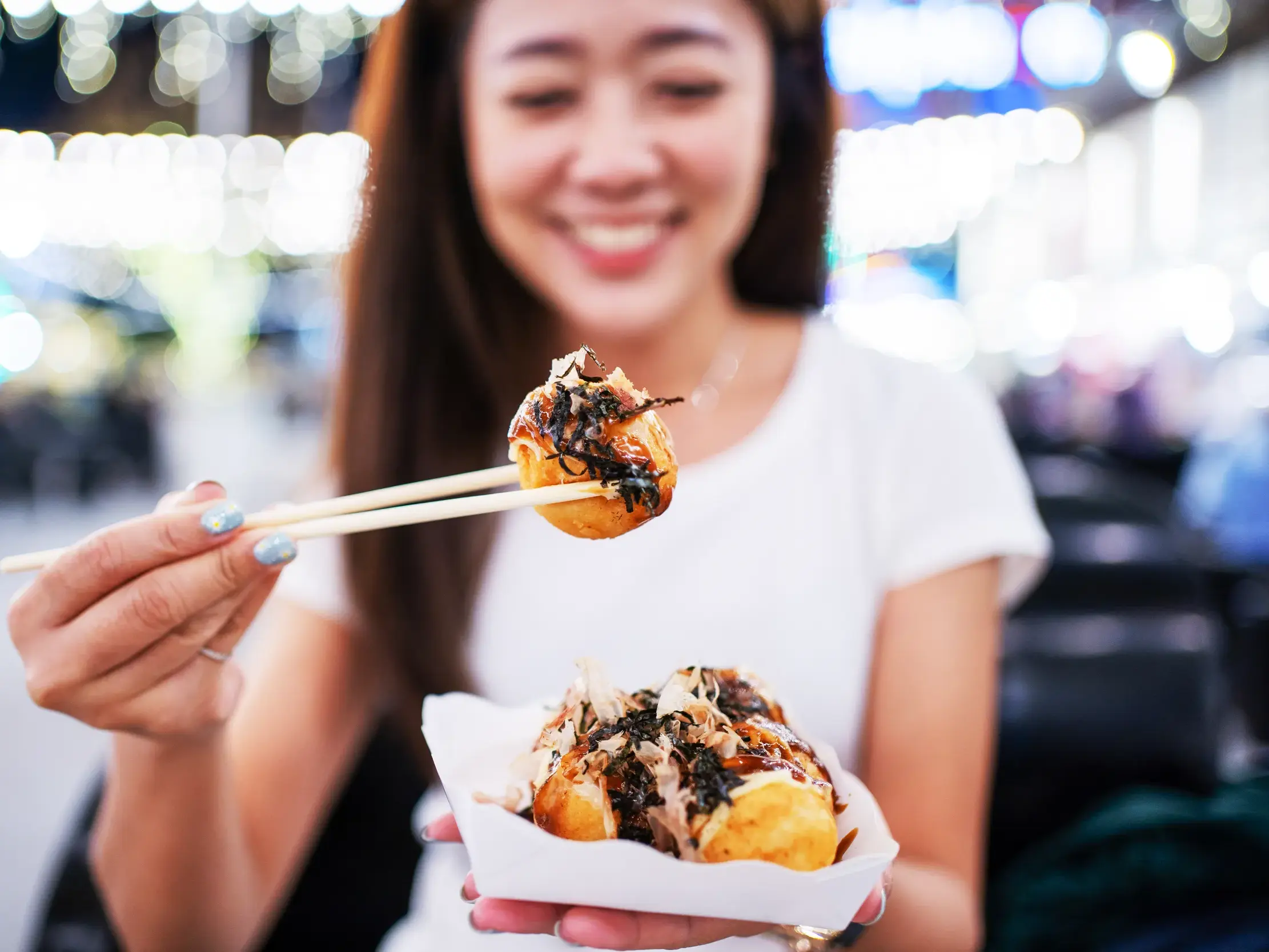 Eine lächelnde Frau hält mit Stäbchen eine Streetfood-Spezialität auf einem bunten Nachtmarkt in Bangkok in die Kamera.