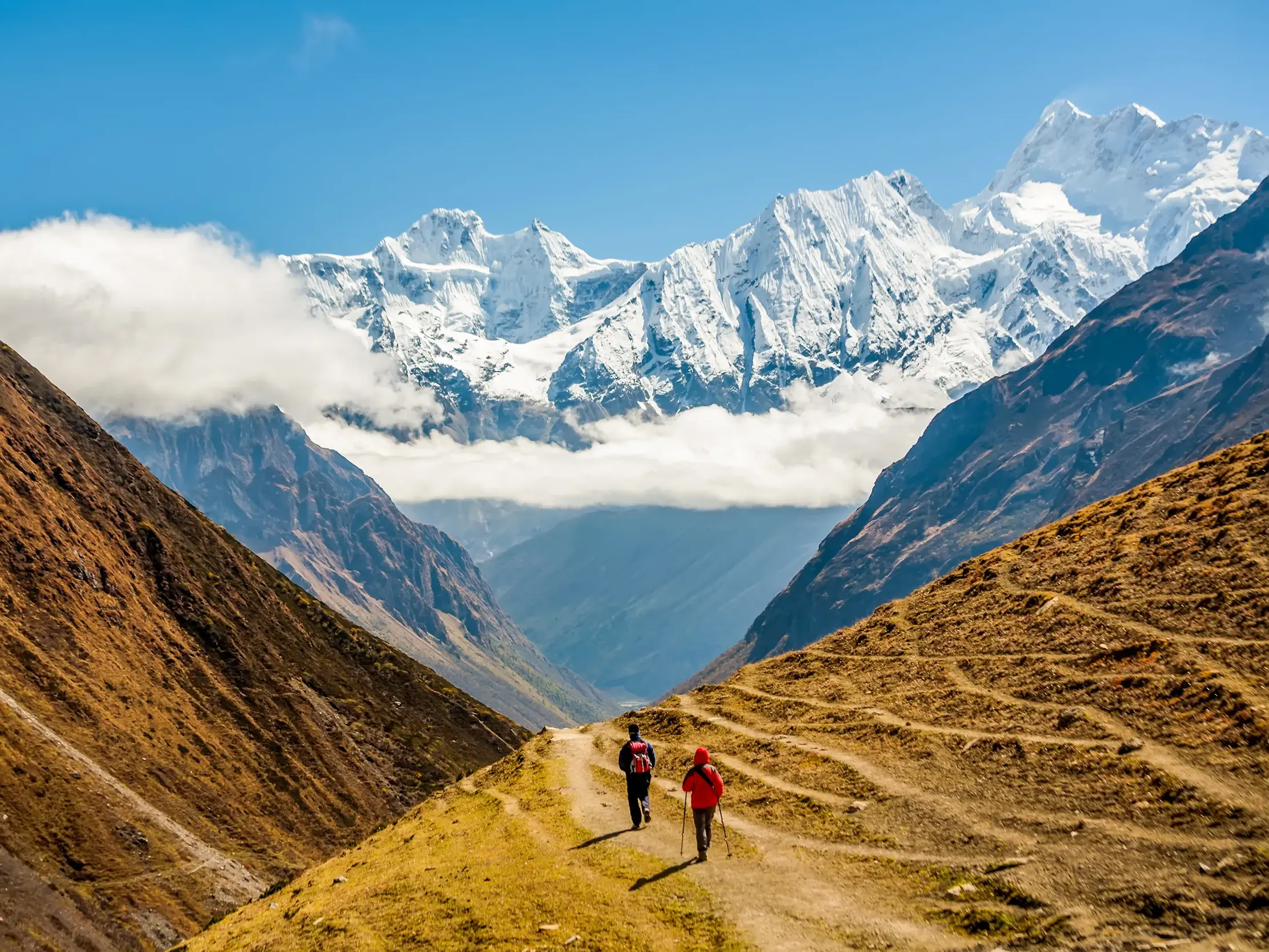 Zwei Wanderer auf einem schmalen Pfad in einem weiten Tal mit Panorama auf die schneebedeckten Gipfel des Manaslu in Nepal.