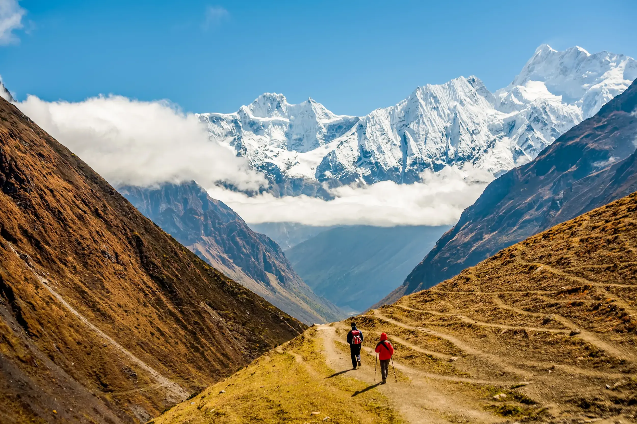 Zwei Wanderer auf einem schmalen Pfad in einem weiten Tal mit Panorama auf die schneebedeckten Gipfel des Manaslu in Nepal.