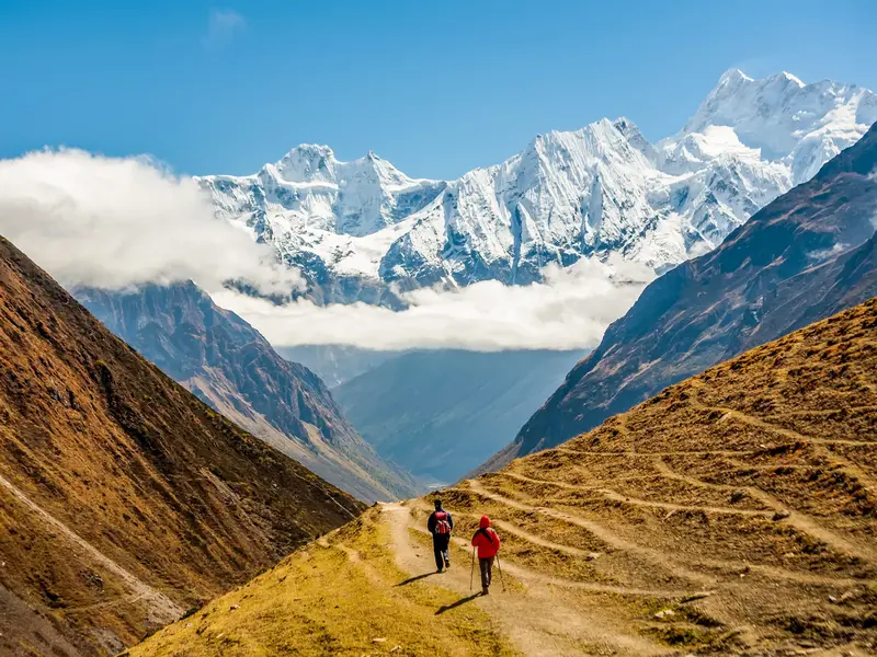 Zwei Wanderer auf einem schmalen Pfad in einem weiten Tal mit Panorama auf die schneebedeckten Gipfel des Manaslu in Nepal.