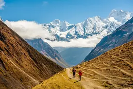 Zwei Wanderer auf einem schmalen Pfad in einem weiten Tal mit Panorama auf die schneebedeckten Gipfel des Manaslu in Nepal.