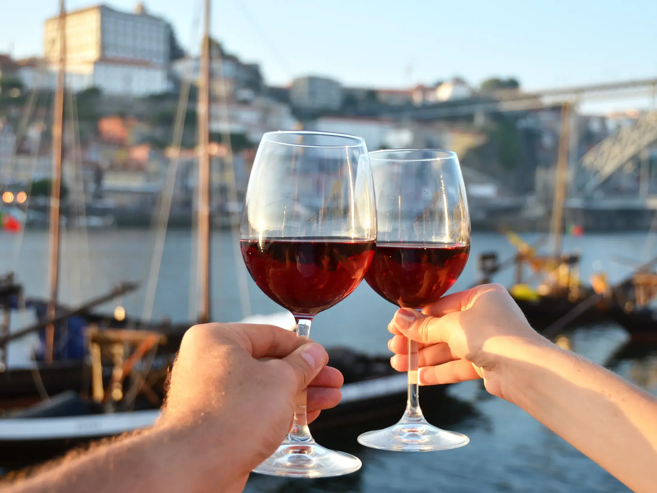 Zwei Personen stoßen mit Rotweingläsern an, im Hintergrund die Portweinboote auf dem Douro im Hafenviertel Ribeira in Porto.
