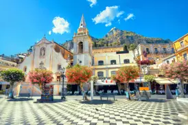 Die sonnige Piazza IX Aprile in Taormina mit der Barockkirche San Giuseppe und Blick auf den Monte Tauro.