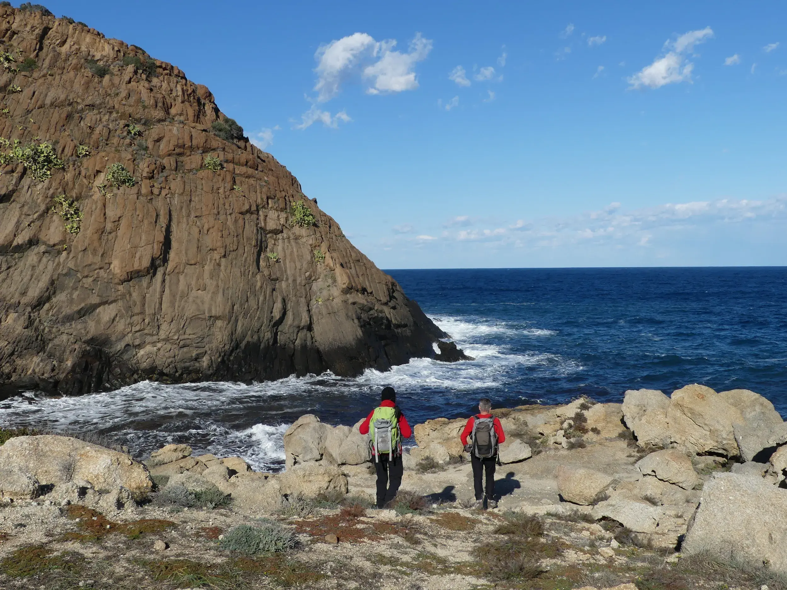 Zwei Wanderer mit Rucksäcken erkunden die felsige Küste am Capo Pecora auf Sardinien mit Blick auf das tiefblaue Meer.
