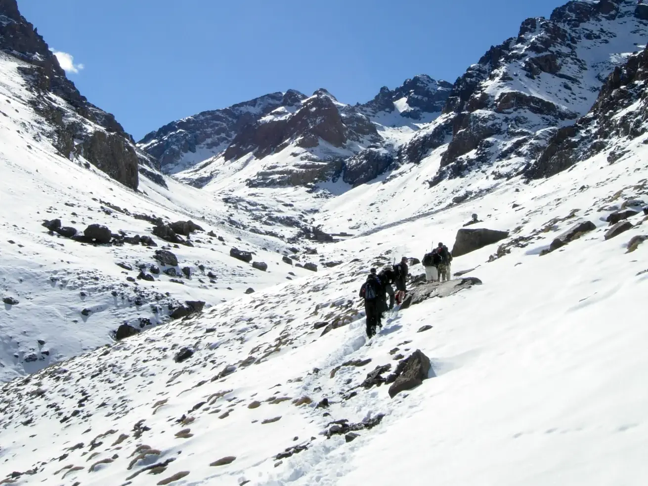 Eine Trekking-Gruppe folgt einem schmalen Pfad durch ein sonniges, schneebedecktes Tal im Hohen Atlas in Marokko.