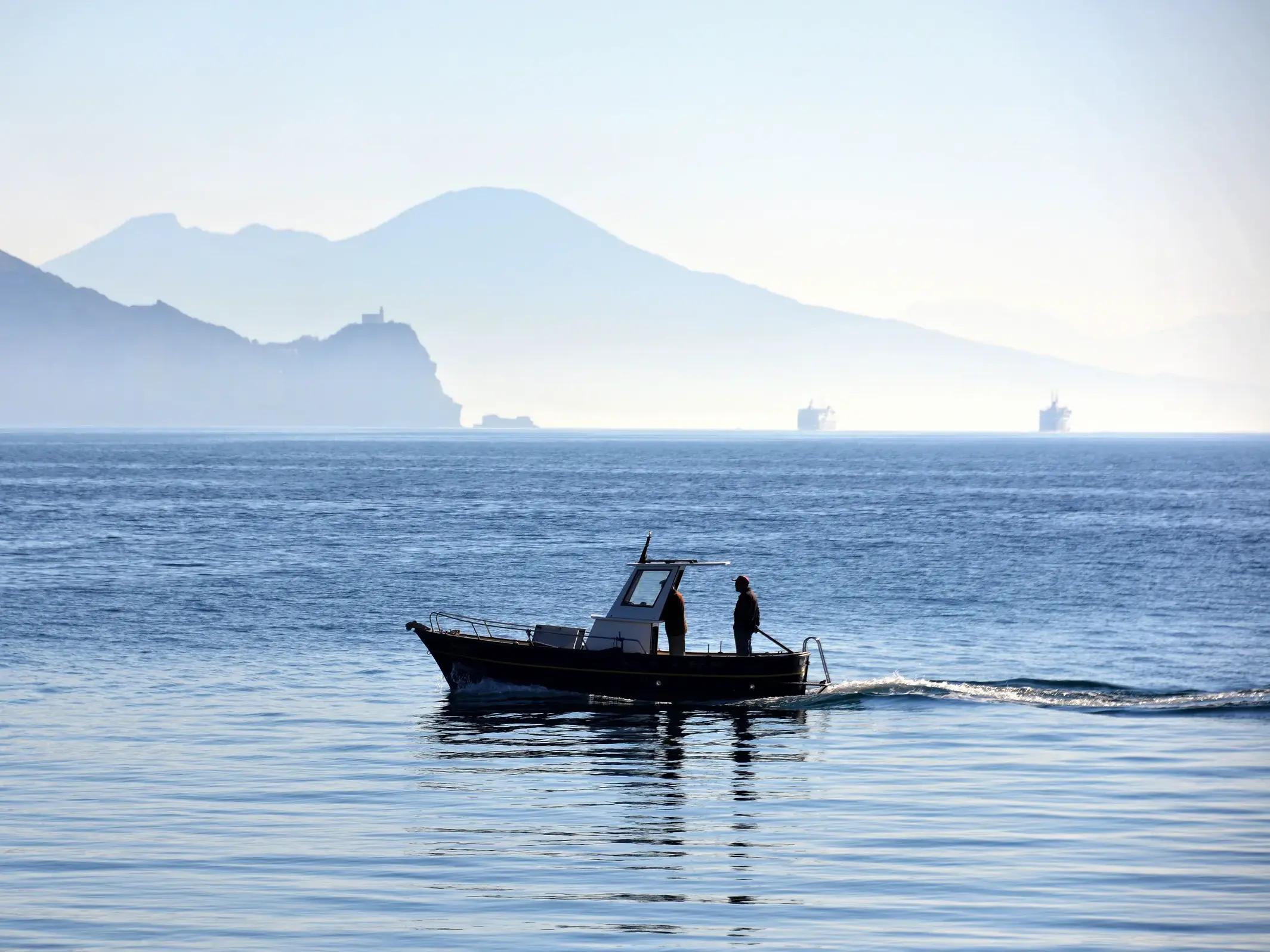 Kleines Motorboot mit zwei Personen bei einer Fahrt auf dem Golf von Neapel vor der Silhouette des Vesuvs im Morgennebel.