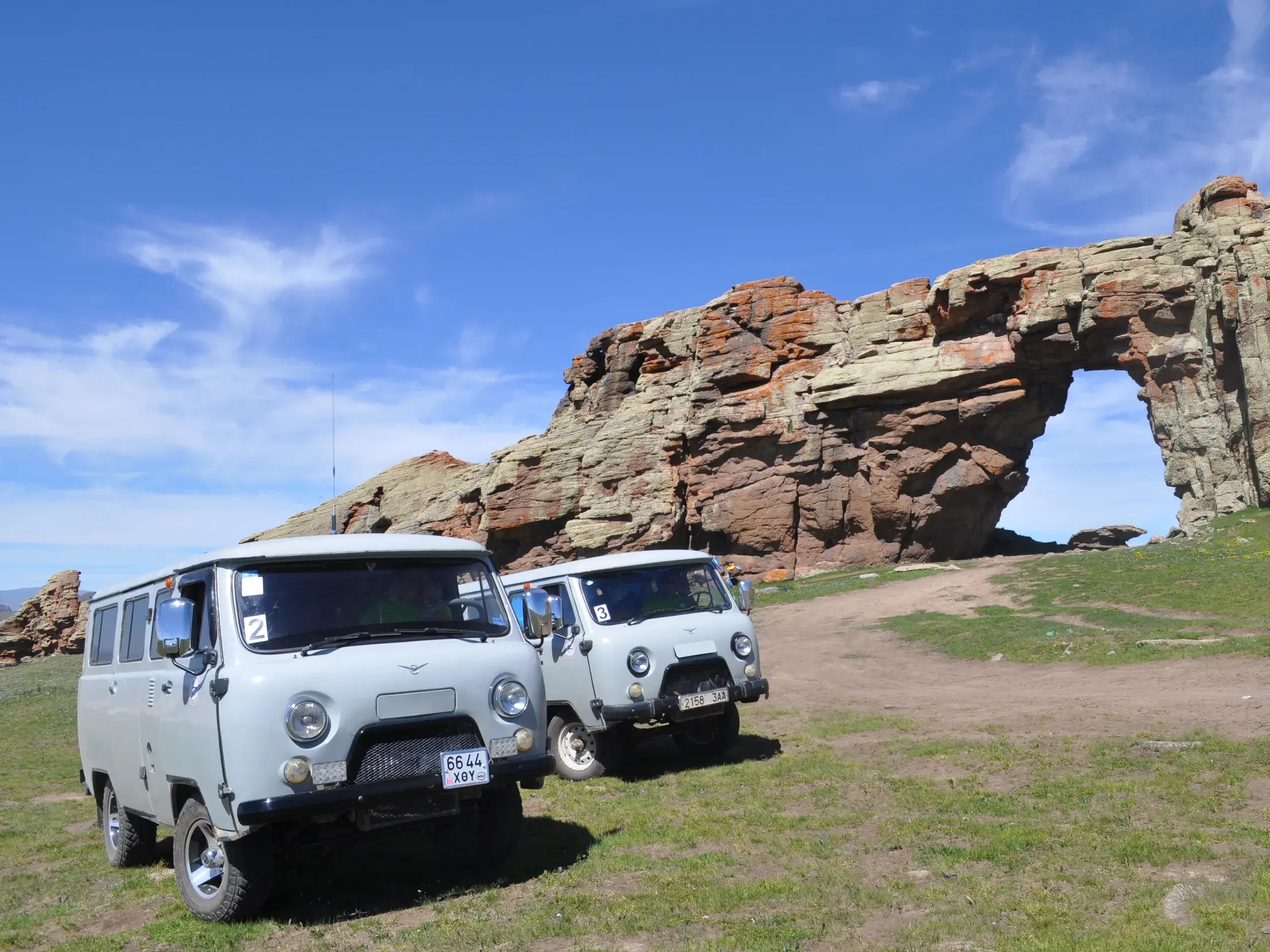 Zwei graue Offroad-Busse parken auf einer Wiese vor dem imposanten, roten Felsentor von Senjit in der Mongolei.