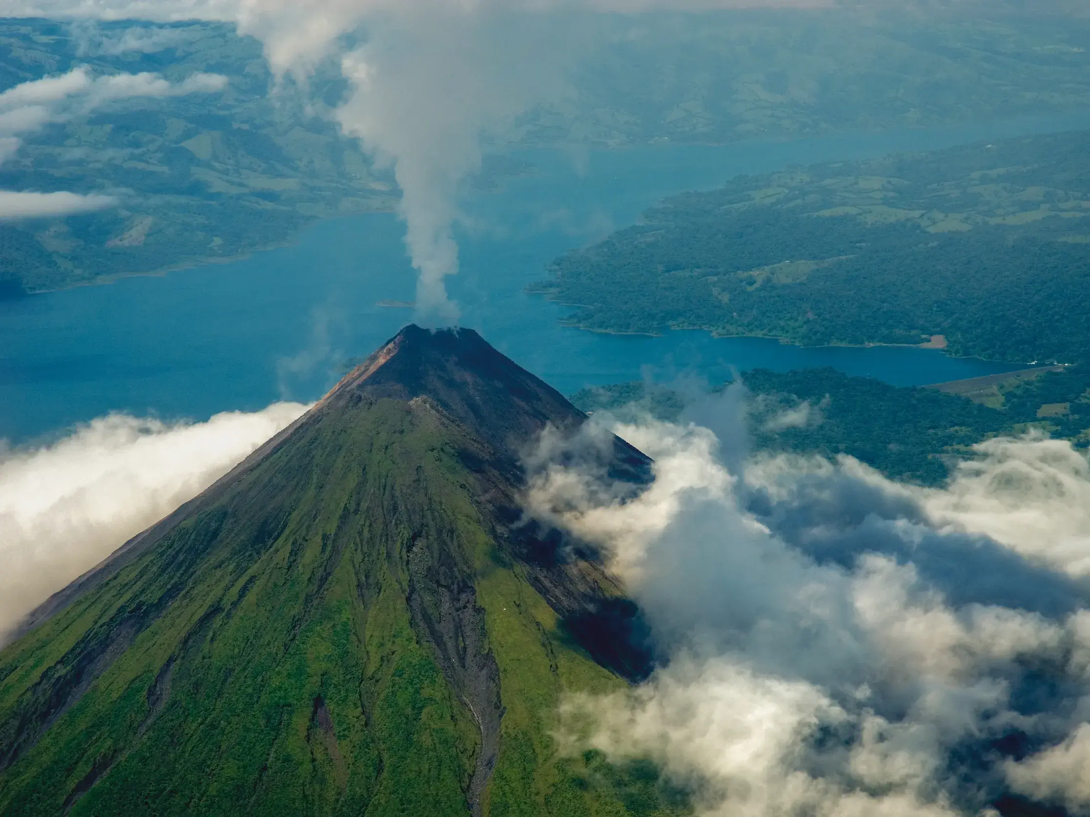 Luftaufnahme des rauchenden Vulkans Arenal in Costa Rica, umgeben von Wolken über dem Arenalsee und grüner Landschaft.