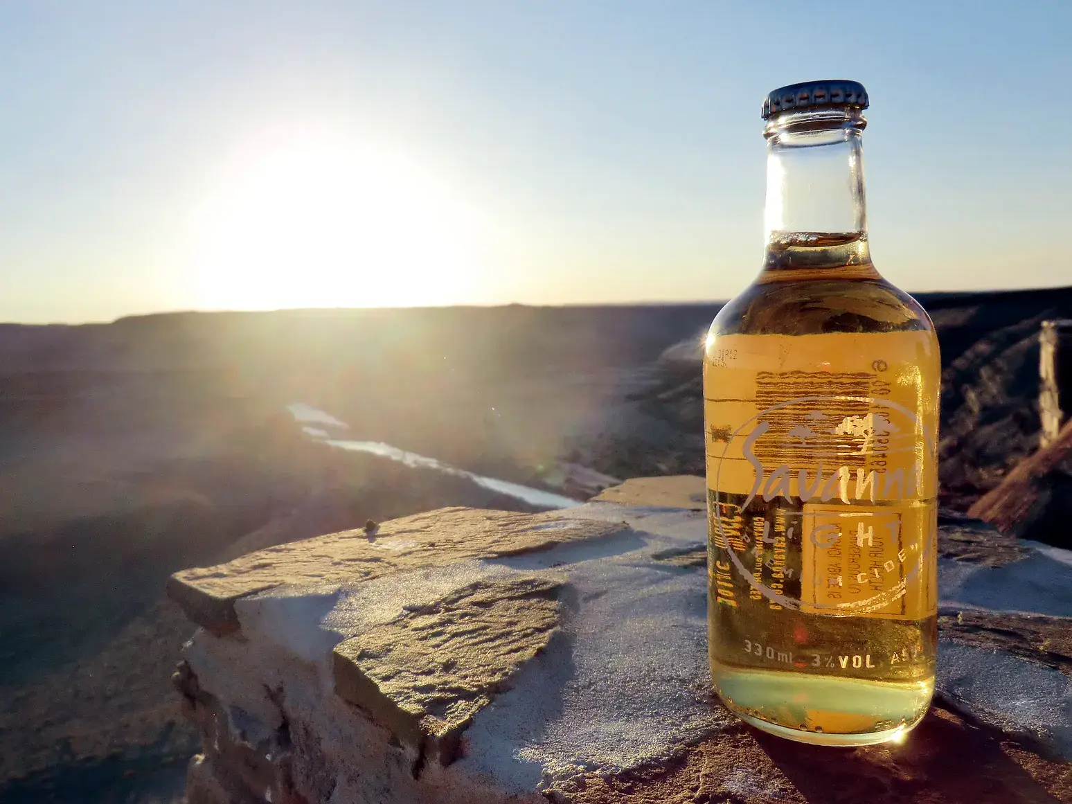 Ein kühles Getränk steht für einen Sundowner auf einem Felsen mit Blick auf den weiten Fish River Canyon in Namibia bei Sonnenuntergang.