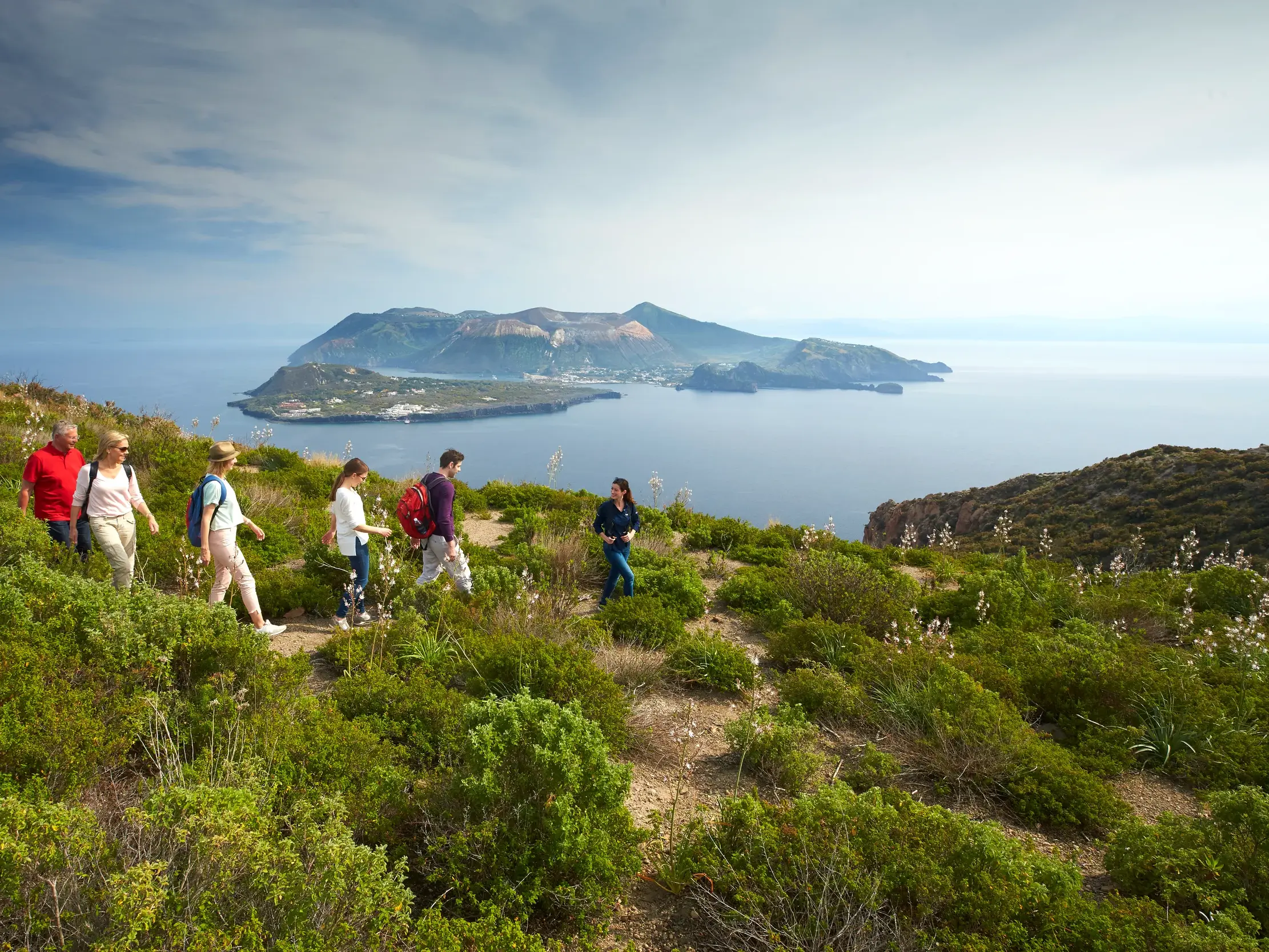 Eine kleine Reisegruppe wandert auf einem grünen Küstenpfad in Sizilien mit weitem Blick über das Meer auf eine Insel.