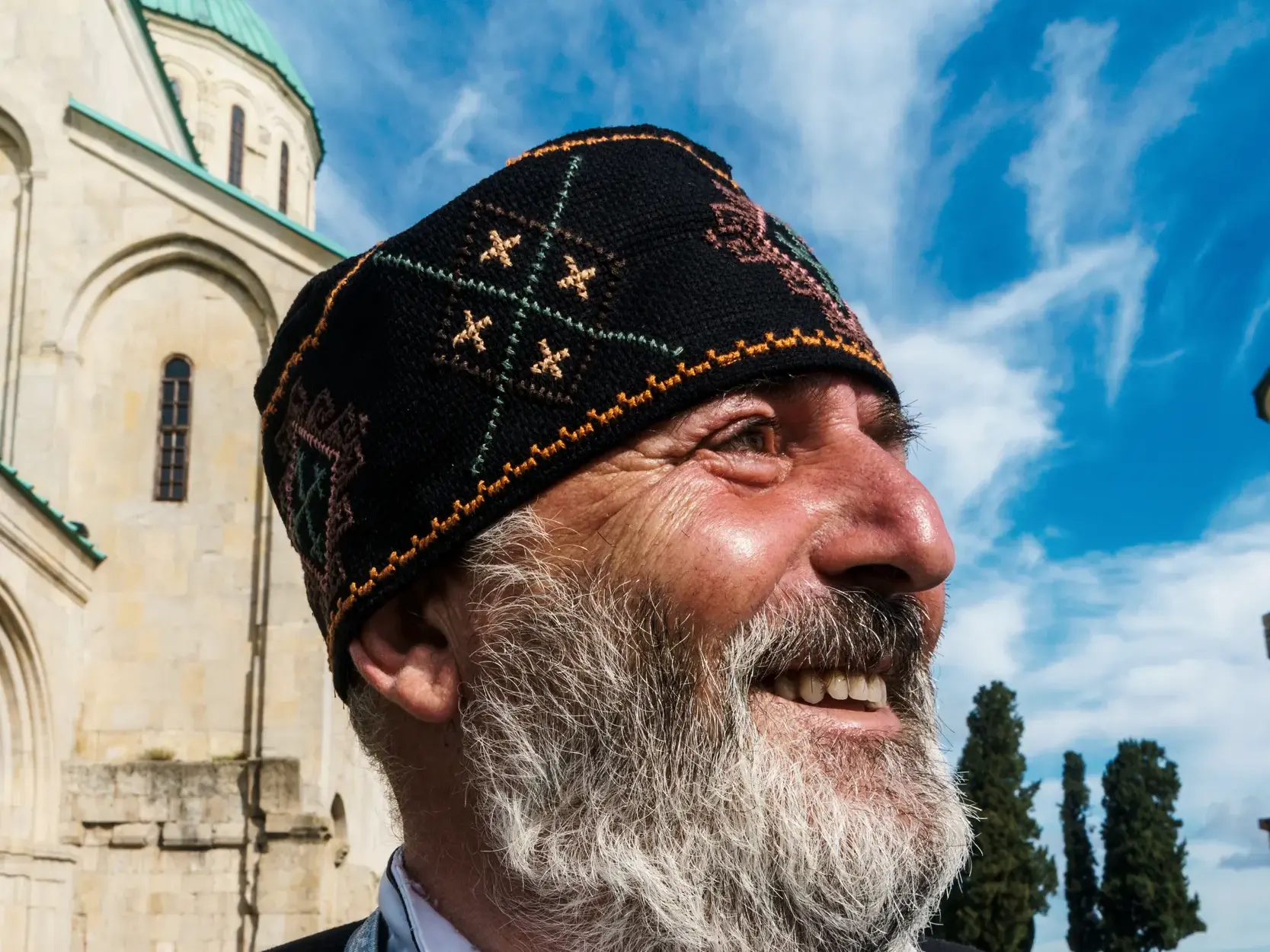 Nahaufnahme eines freundlichen Mannes mit Bart und bestickter Mütze, der vor einer historischen Kirche in Georgien lächelt.
