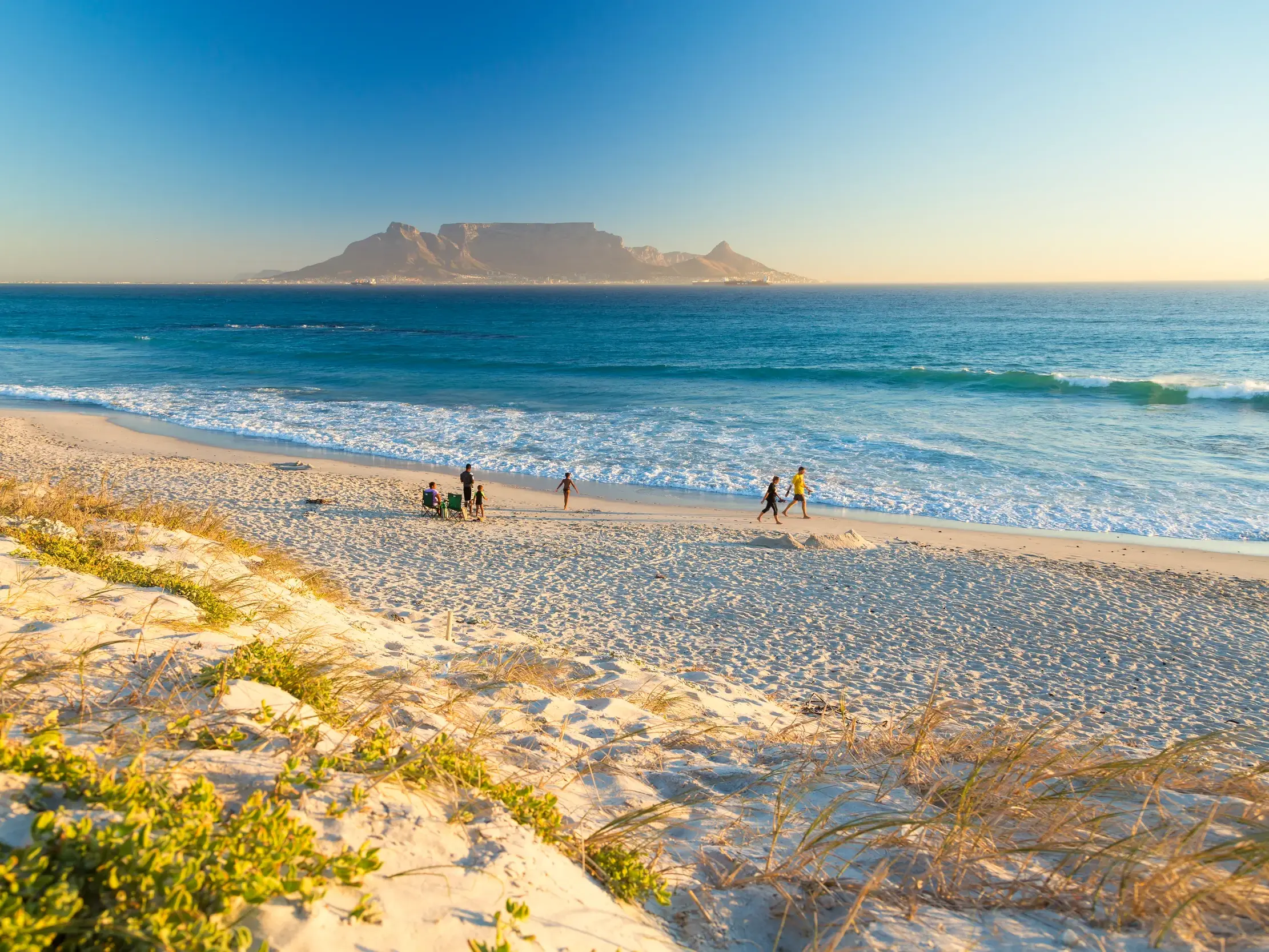 Weite Dünenlandschaft an einem Sandstrand bei Kapstadt mit Blick auf den berühmten Tafelberg in der Abendsonne.