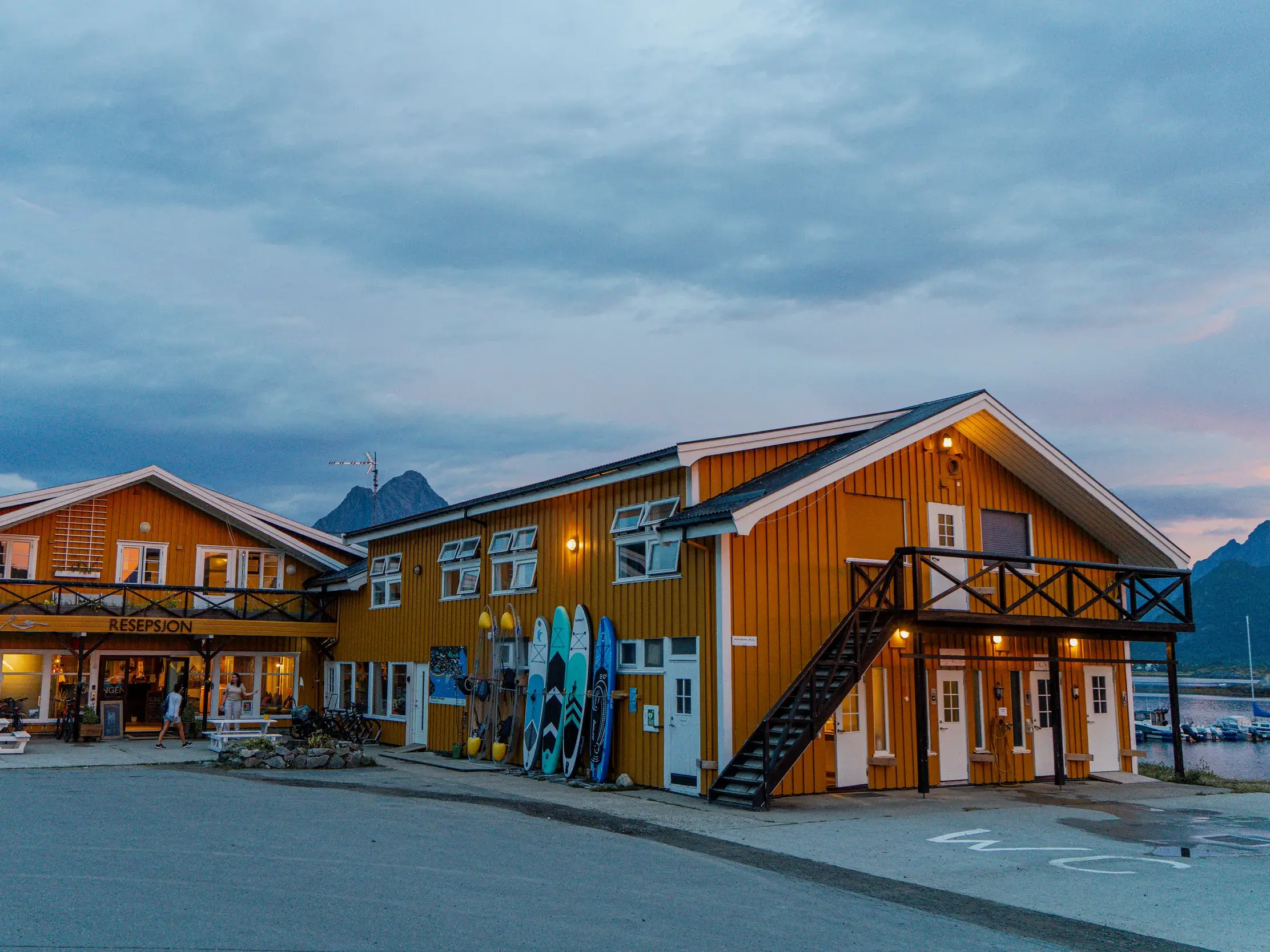 Zwei beleuchtete, gelbe Hotelgebäude im traditionellen Stil am Hafen auf den Lofoten vor einer Bergkulisse bei Dämmerung.