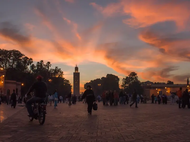 Abenddämmerung in Marrakesch: Menschen flanieren auf einem belebten Platz vor der Koutoubia-Moschee unter orangenem Himmel.