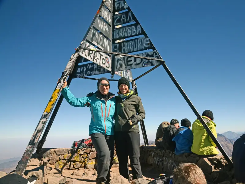 Zwei glückliche Wanderinnen feiern ihre erfolgreiche Trekking-Tour am Gipfel des Djebel Toubkal mit der markanten Pyramide.
