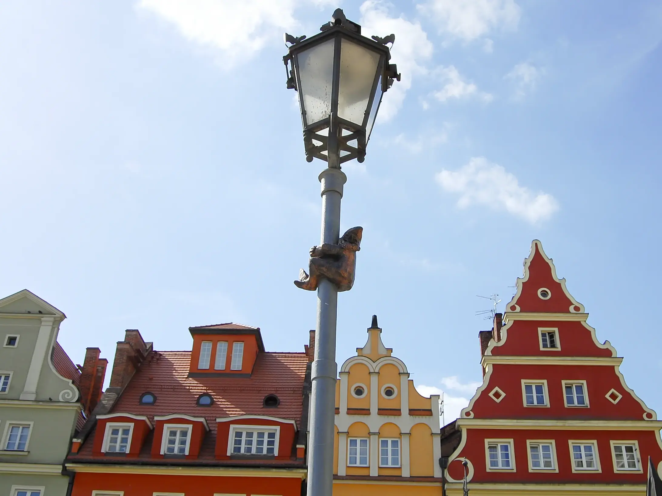 Bronzene Zwergfigur auf Entdeckungstour an einem Laternenpfahl vor der bunten Häuserfassade des Rings in Breslau.