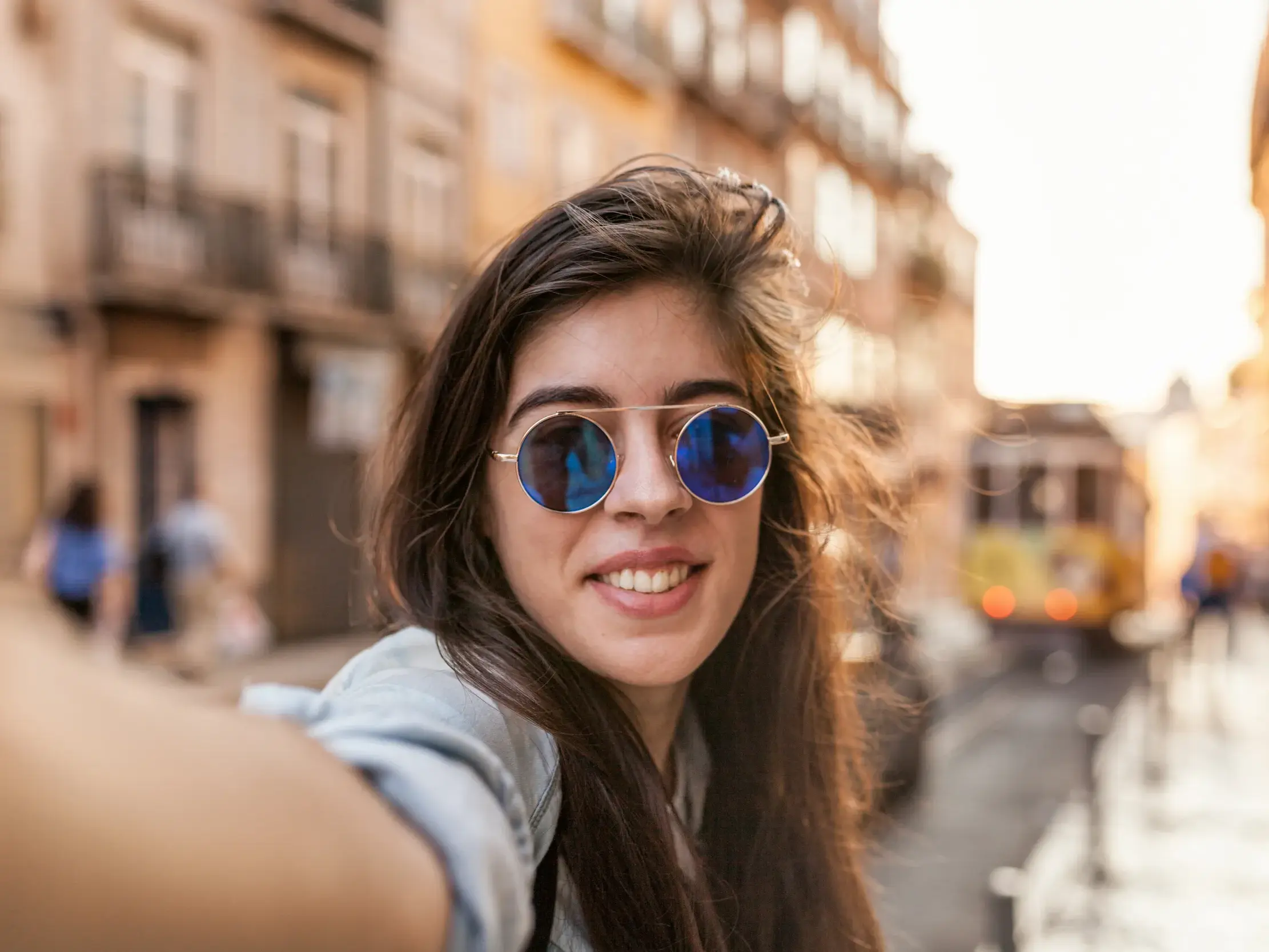 Junge Frau auf Entdeckertour macht ein Selfie in einer Gasse in Lissabon, im Hintergrund die ikonische gelbe Straßenbahn.