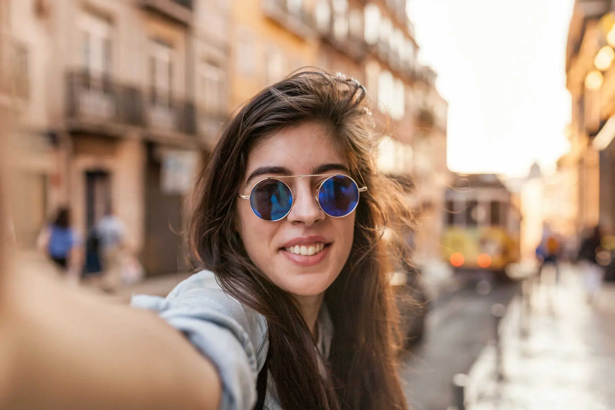 Junge Frau auf Entdeckertour macht ein Selfie in einer Gasse in Lissabon, im Hintergrund die ikonische gelbe Straßenbahn.