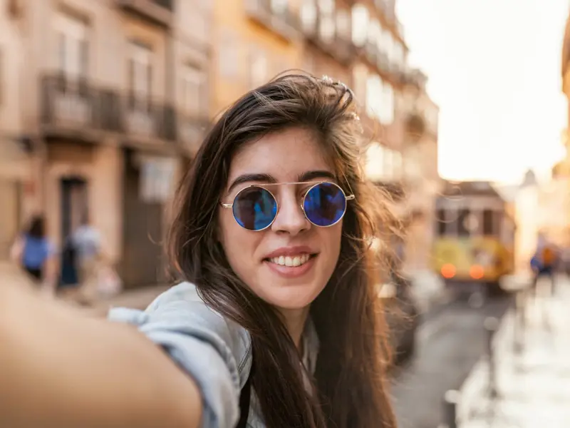 Junge Frau auf Entdeckertour macht ein Selfie in einer Gasse in Lissabon, im Hintergrund die ikonische gelbe Straßenbahn.