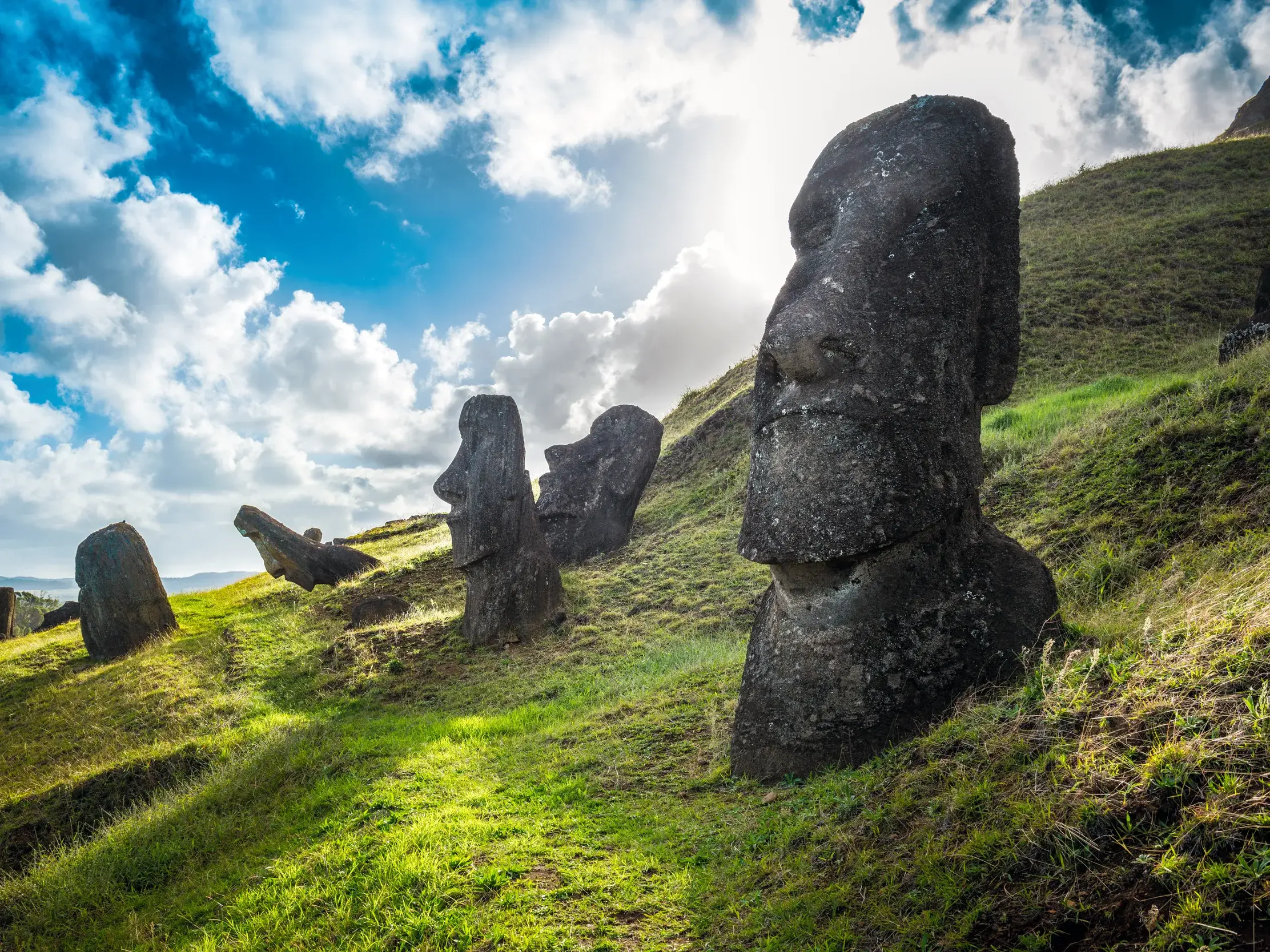 Mystische Moai-Statuen am grasbewachsenen Hang des Vulkans Rano Raraku auf der Osterinsel unter dramatischem Himmel.