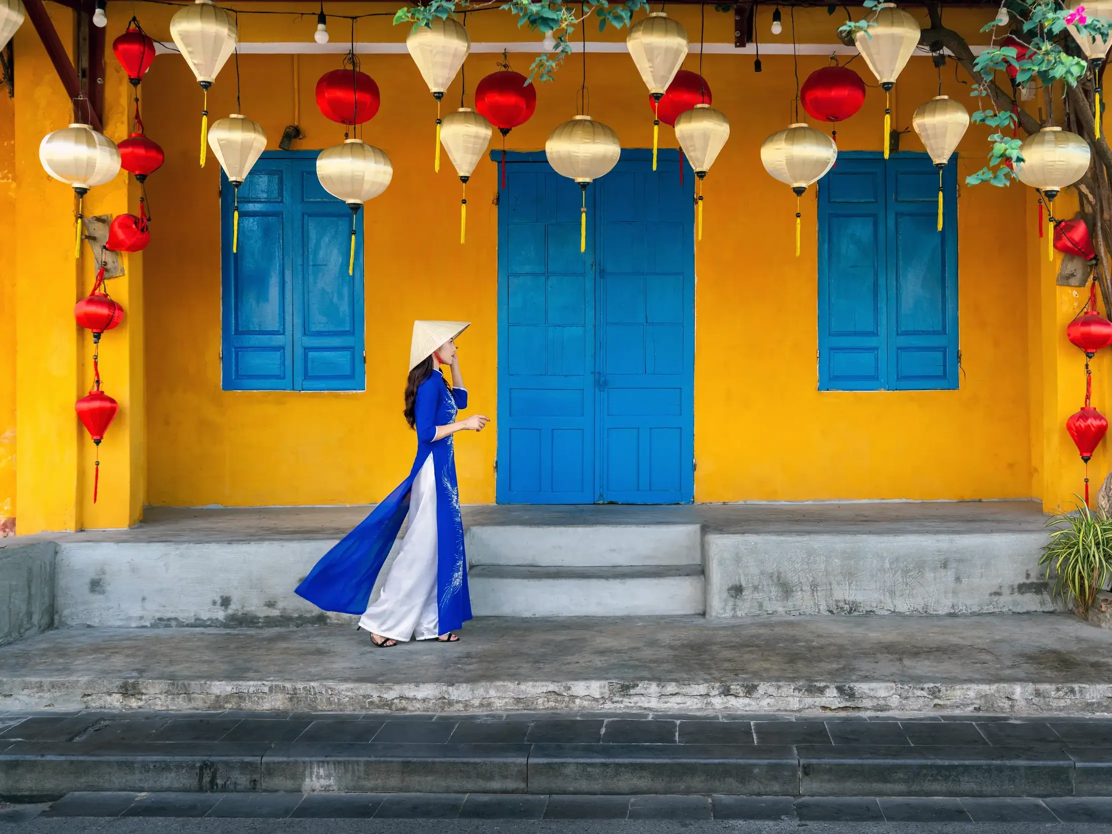 Vietnamesin in blauem Áo dài und Nón-Lá-Hut vor einer gelben Hausfassade mit Lampions in der Altstadt von Hoi An in Vietnam.