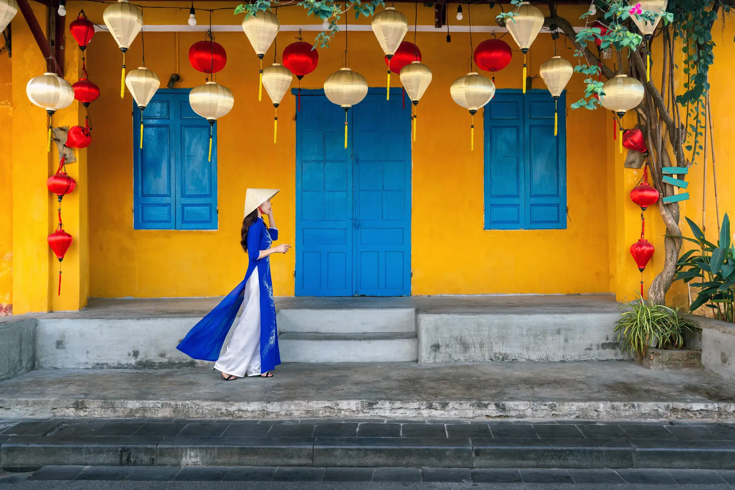 Vietnamesin in blauem Áo dài und Nón-Lá-Hut vor einer gelben Hausfassade mit Lampions in der Altstadt von Hoi An in Vietnam.