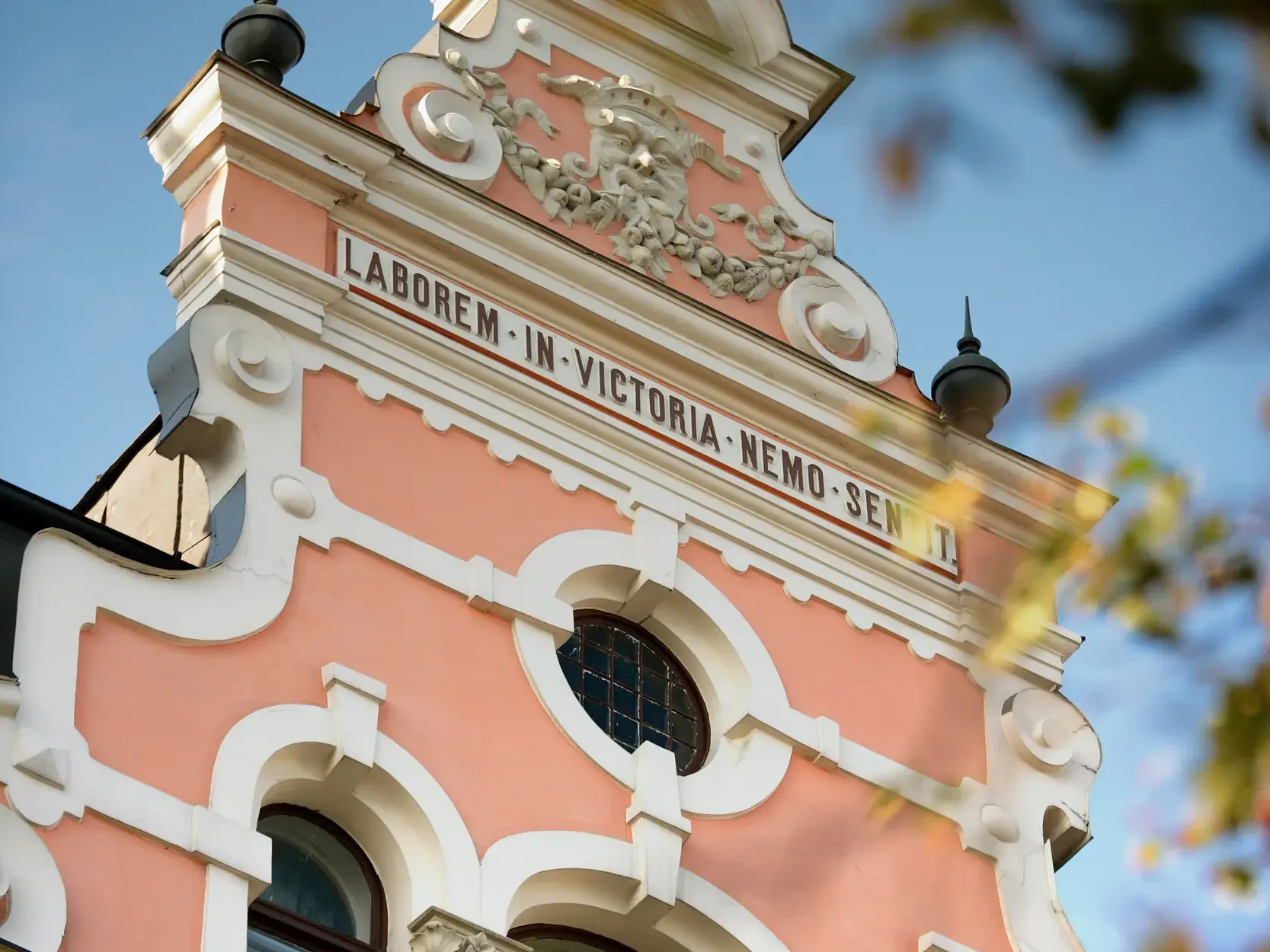Reich verzierter Giebel einer rosa-weißen Barockfassade mit lateinischer Inschrift in der Altstadt von Vilnius.